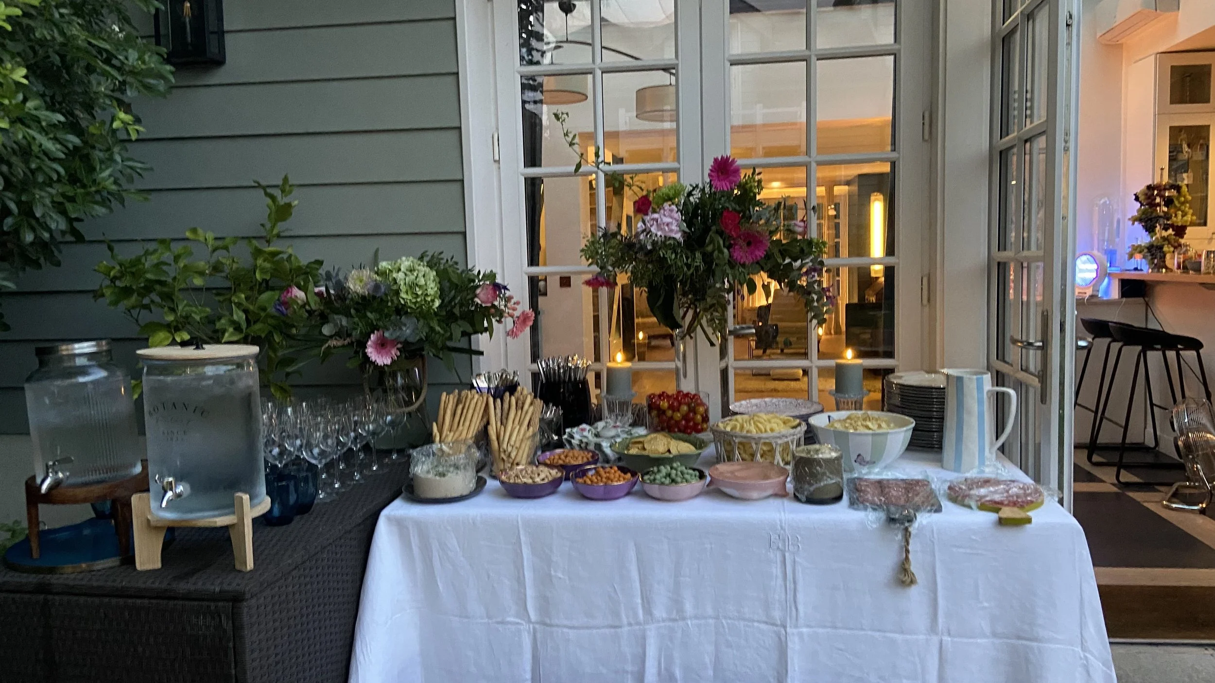Table dressée avec divers mets et boissons dans un jardin d'Ile de France avec grande porte-fenêtre donnant sur un intérieur chaleureux.