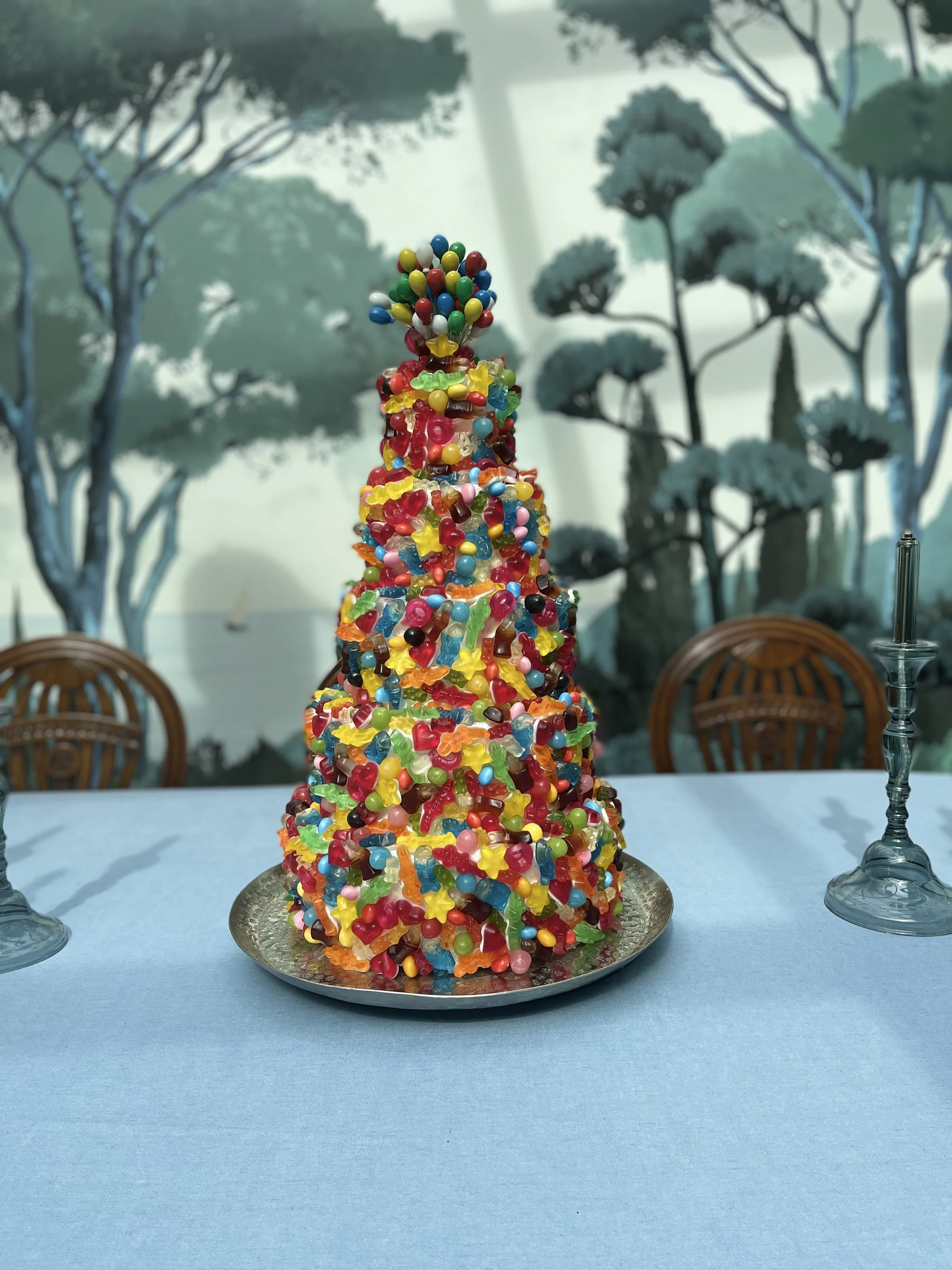 Gâteau en forme de tour, recouvert de bonbons colorés, placé sur un plateau d'argent, avec des verres à vin sur la table et un décor arboré en arrière-plan.