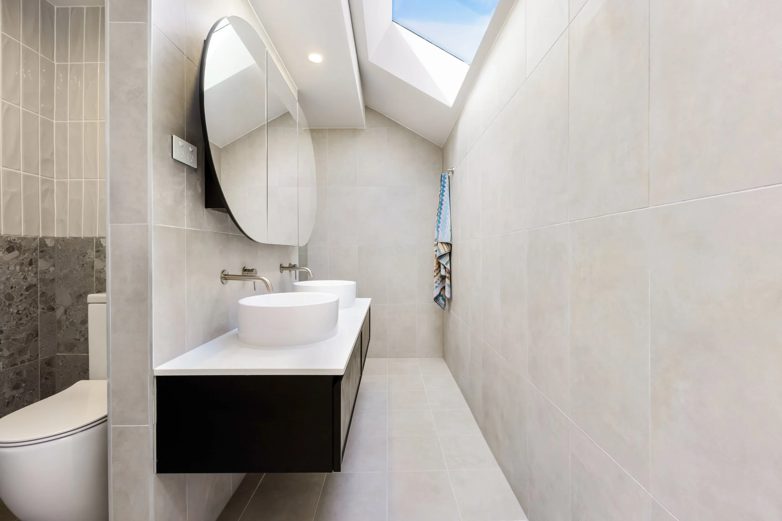 Modern bathroom with a skylight, double vessel sinks, and a large mirror above a dark vanity, with light-colored tiled walls and floor. Sydney Park Terraces
