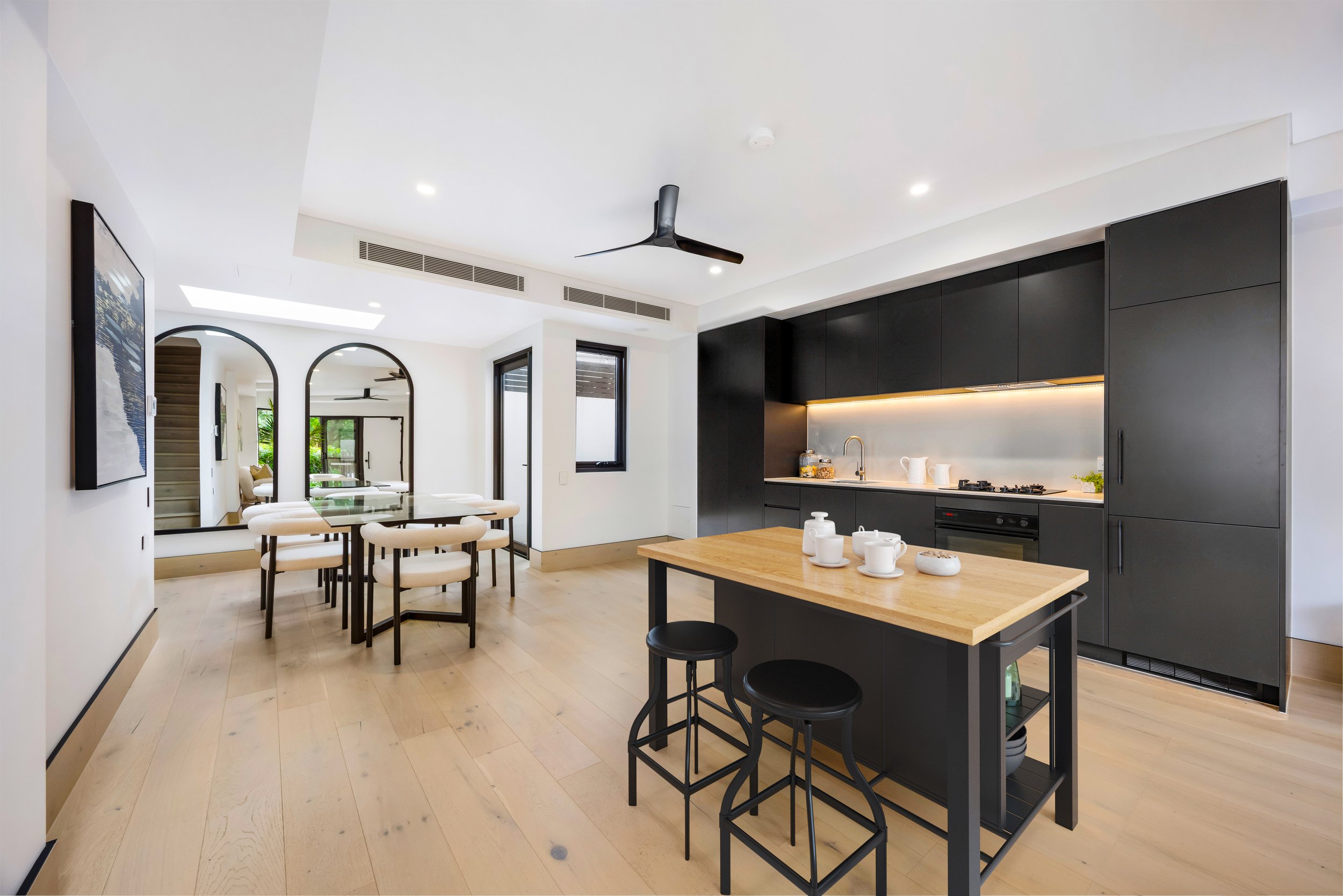 Modern kitchen and dining area with black cabinetry, white walls, light wood flooring, and a wooden island with black stools. Dining table with beige chairs and round mirror accents on the wall. Sydney Park Terraces