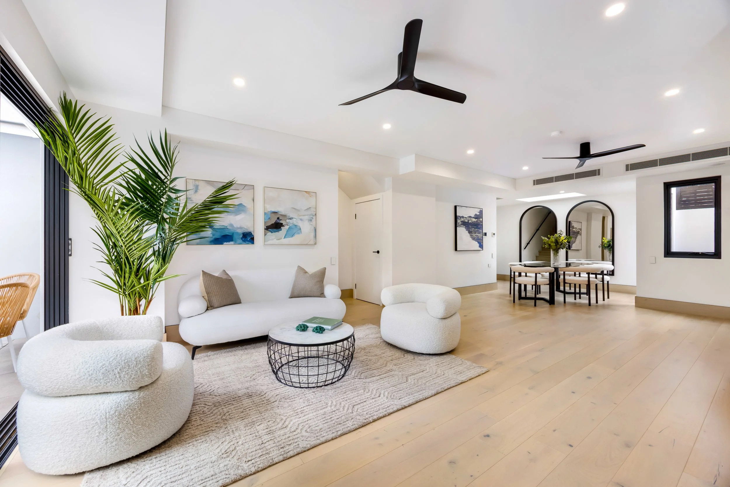 Modern open-concept living room with white walls, light wood flooring, and contemporary furniture including round armchairs, a sofa, and a round coffee table, decorated with abstract art and lush green plants, with black ceiling fans and ceiling ligh