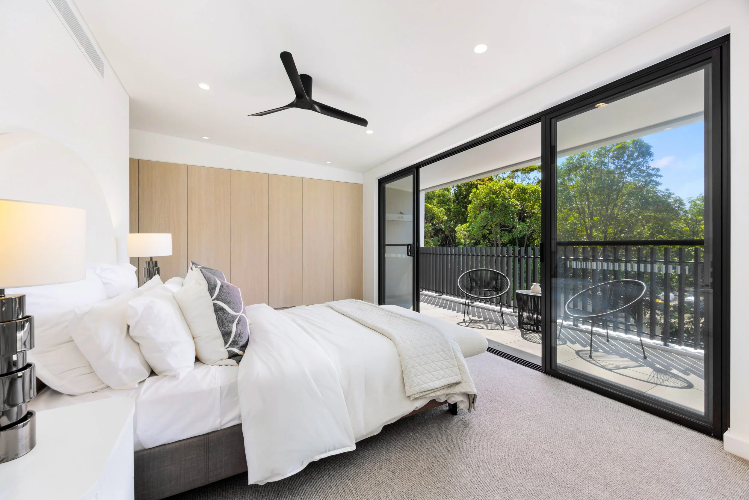 Modern bedroom with a large bed, white bedding, decorative pillows, bedside lamps, a wooden wardrobe, and a glass sliding door leading to a balcony with outdoor seating and greenery view. Sydney Park Terraces