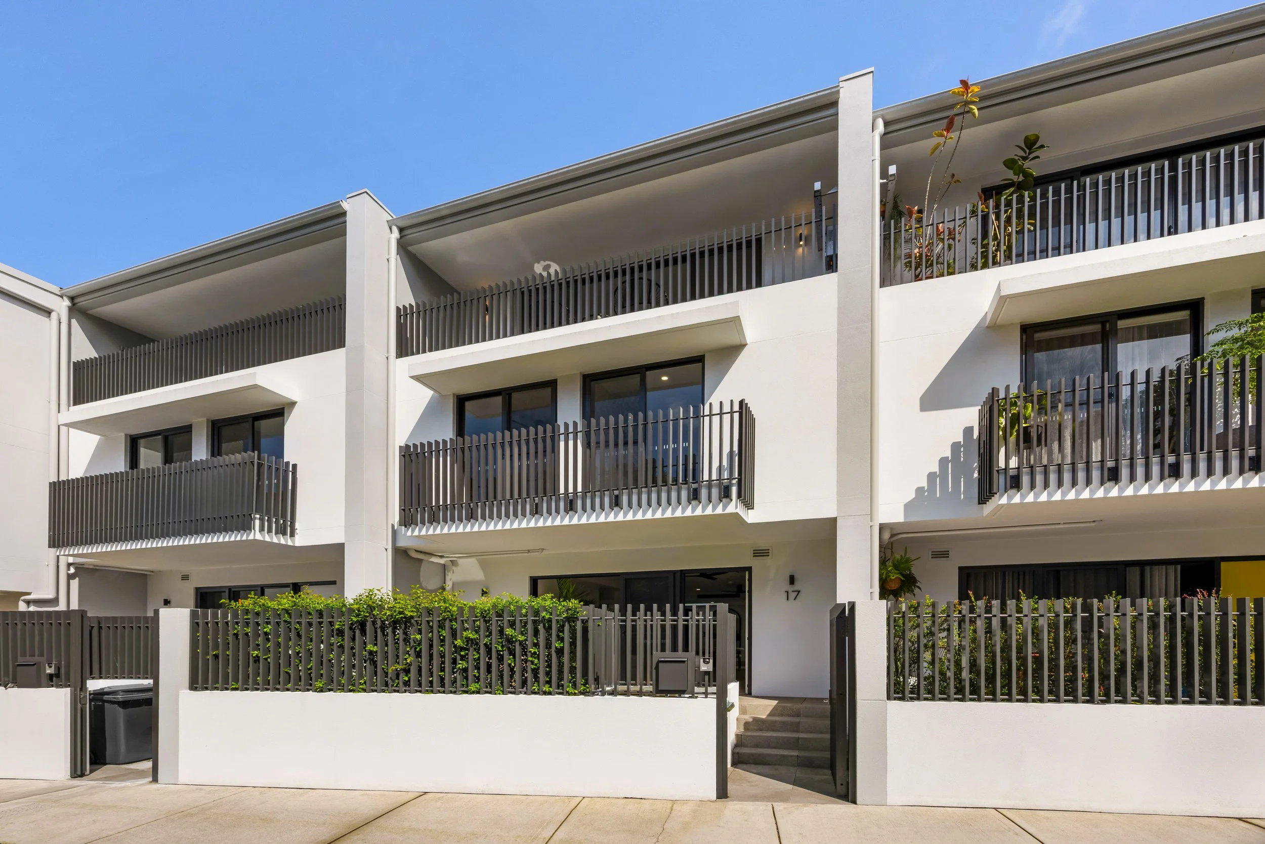 Modern white apartment building with multiple balconies, black railings, and glass doors, surrounded by a sidewalk and greenery, under a clear blue sky. Sydney Park Terraces