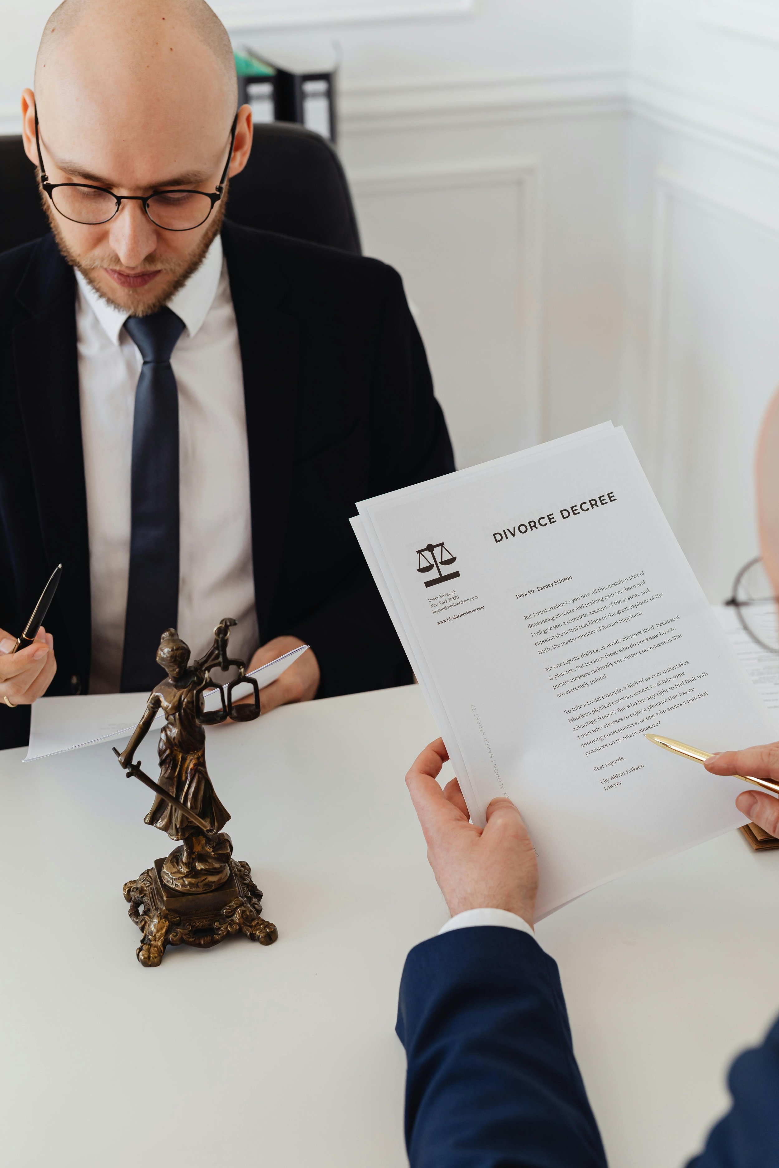 A man in a black suit and glasses is reading a document titled 'Divorce Decree' at a white desk, with a person in a blue suit pointing at another page, and a bronze statue of Lady Justice on the table.