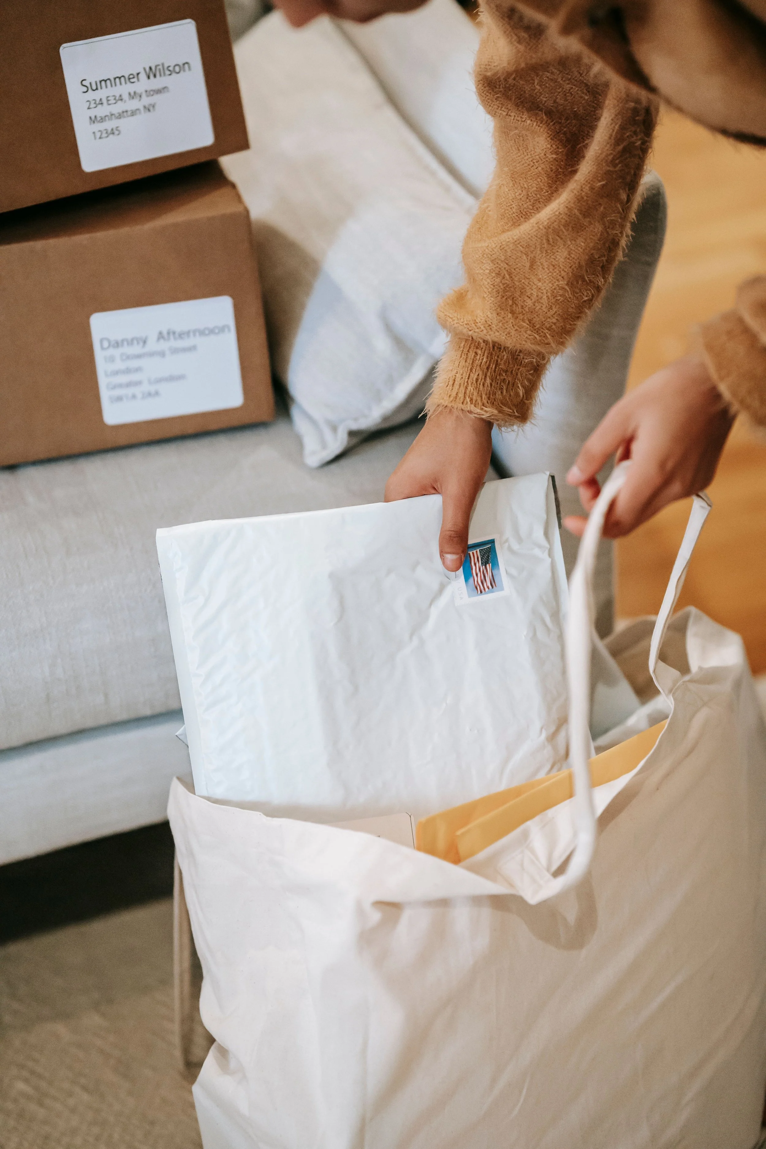 Person packing white envelopes into a paper bag, with cardboard boxes labeled with names and addresses in the background.