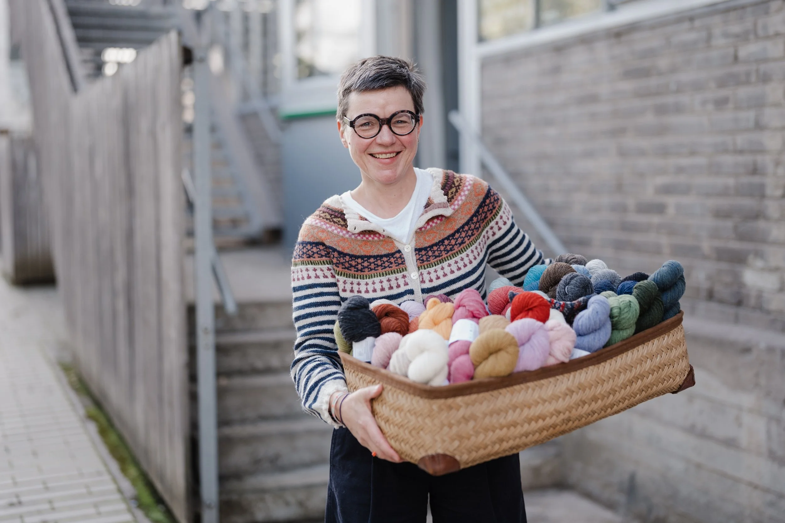 Une femme souriante portant des lunettes, tenant un grand panier rempli de fils à tricoter de différentes couleurs, devant un mur en briques et un cluaire à l'extérieur.