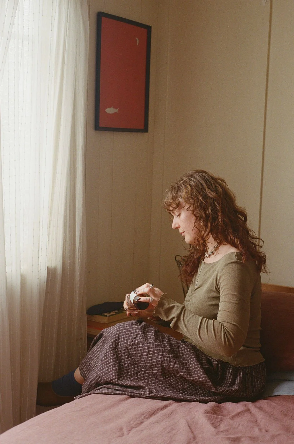 A young woman with curly red hair sitting on a bed, looking at a bottle she is holding in her hands, in a softly lit room with beige walls and a framed artwork of a fish and moon on the wall.
