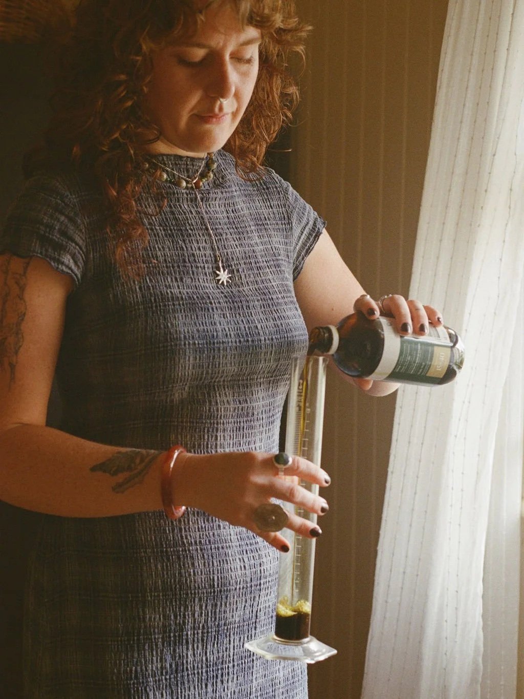 A woman with curly red hair wearing a gray patterned dress, holding a presser apparatus with alcohol and a test tube, standing in front of a window with white curtains.