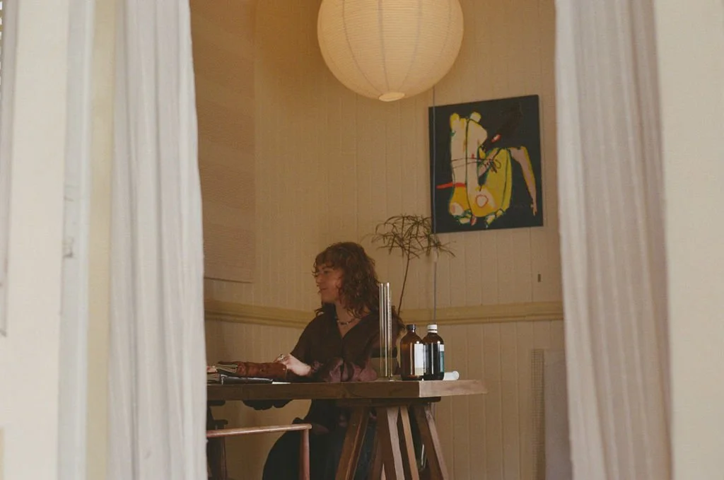 Woman sitting at a wooden table in a room with beige walls, a large pendant lamp, and abstract art on the wall, viewed through a doorway.
