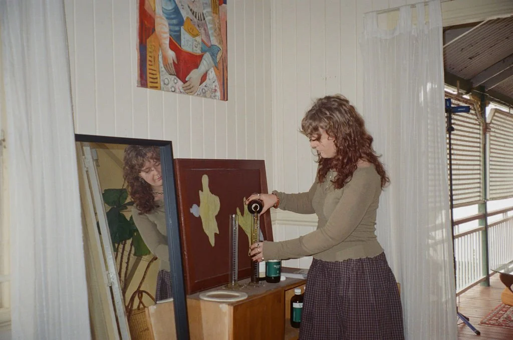 A woman with curly hair and glasses stands in front of a mirror, holding a tape measure, in a room with white paneled walls and a colorful painting above her. She appears to be measuring or adjusting a piece of art or furniture.