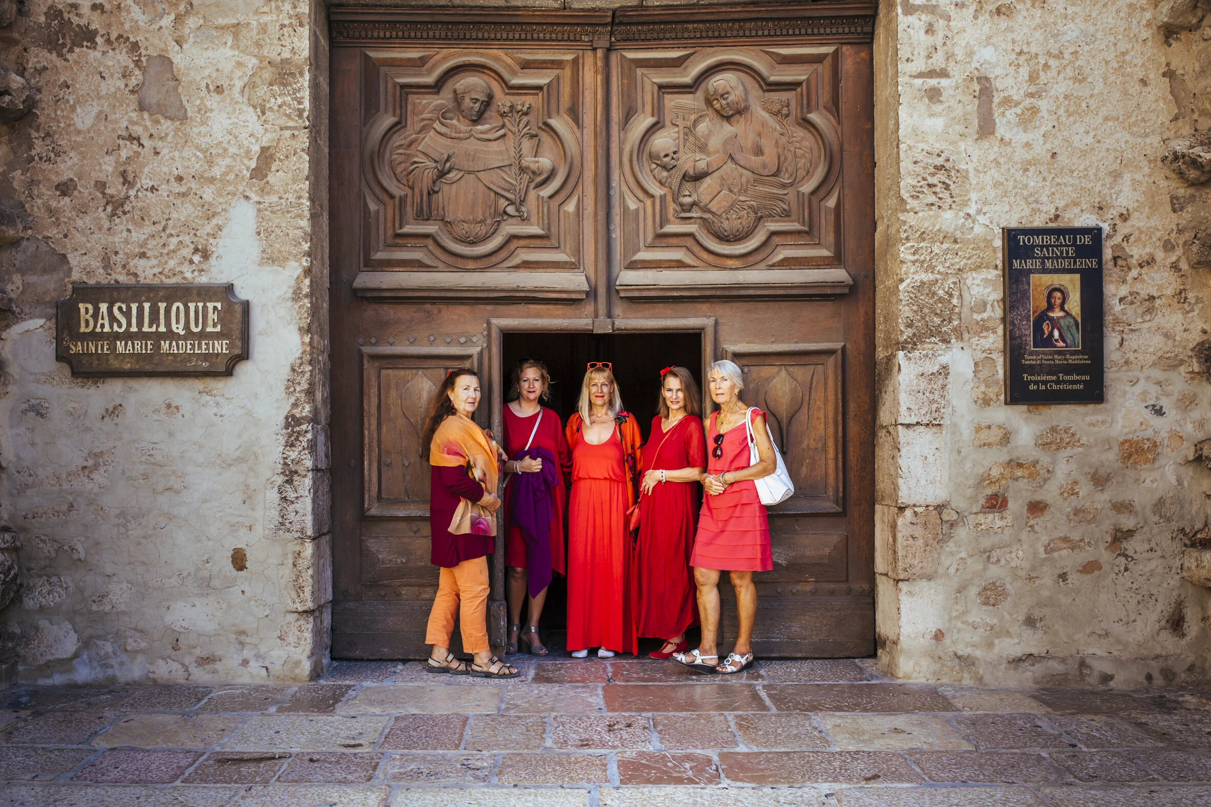 Five women standing together in front of the Saint Mary Madeleine basilica entrance, with carved religious figures and signs on the wall behind them. They are wearing colorful dresses and accessories.