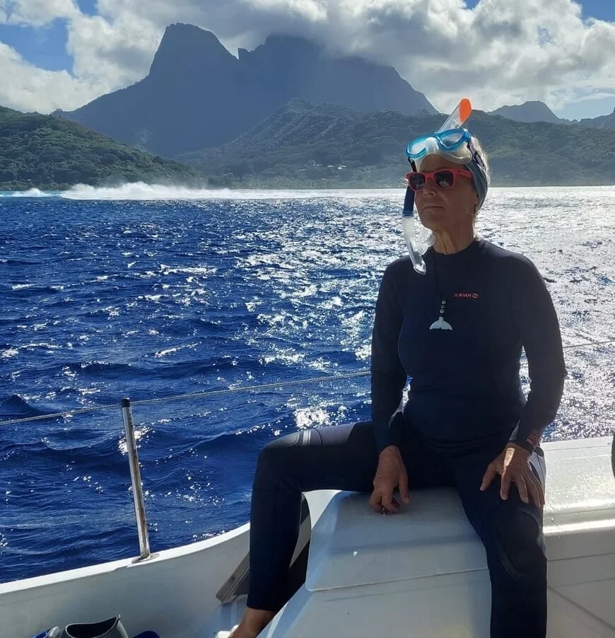Woman with snorkeling gear sitting on a boat with a mountain and ocean in the background.
