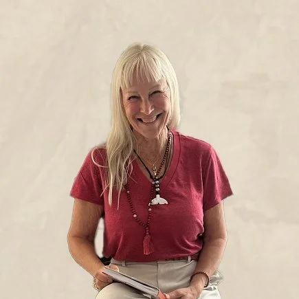A smiling woman with long blonde hair wearing a red shirt, holding a tablet, standing against a plain light-colored background.