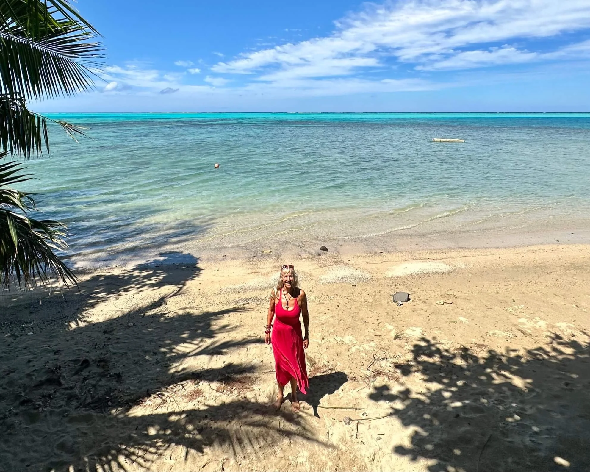 A woman in a pink dress standing on a sandy beach with palm trees, calm ocean waters, and a clear blue sky with clouds in the background.