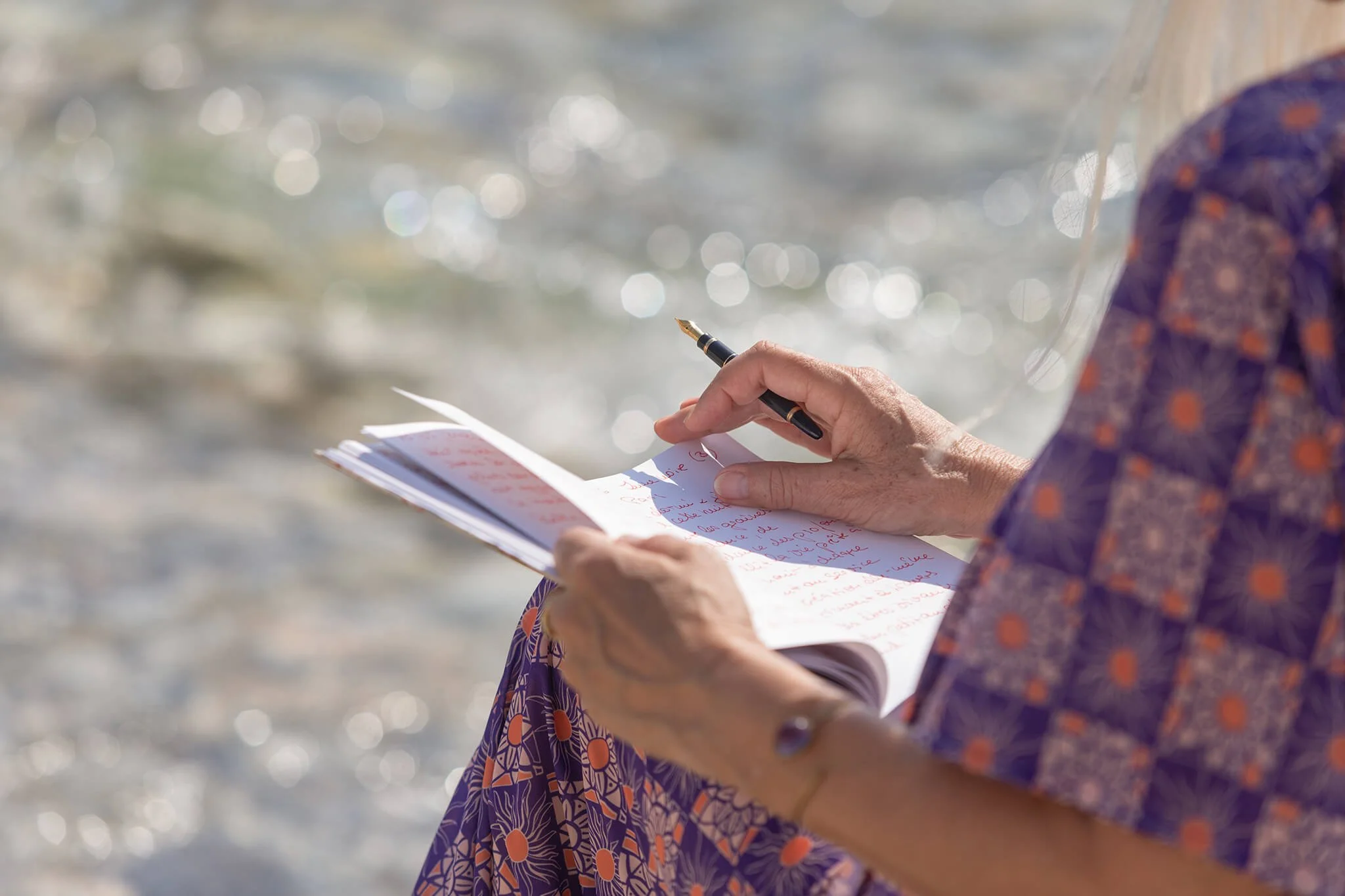 A person in a colorful patterned dress is writing in a notebook with a fountain pen, outdoors near water with sunlight creating bokeh effects in the background.