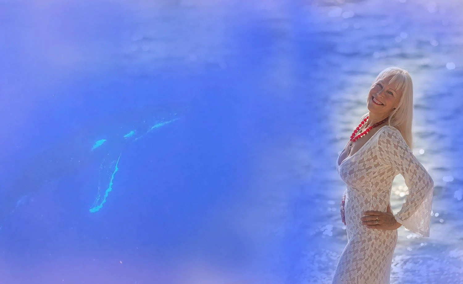 A smiling woman with blonde hair, wearing a white lace dress and a red beaded necklace, stands on the beach near the water with her hands on her hips, and is connected to the whales and the ocean