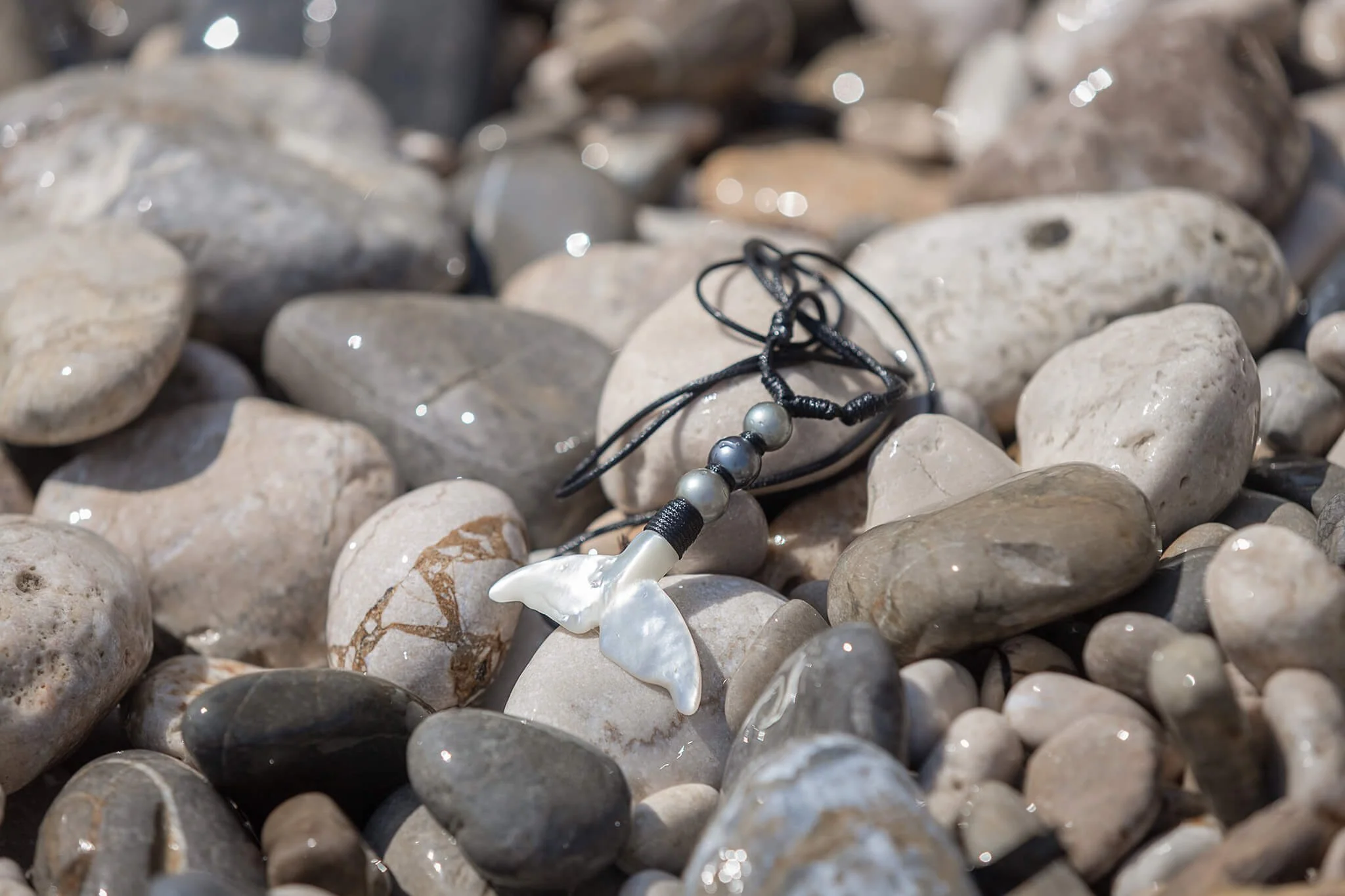 A whale necklace with large grey pearls lying on a bed of smooth, rounded pebbles with some shiny, wet spots.