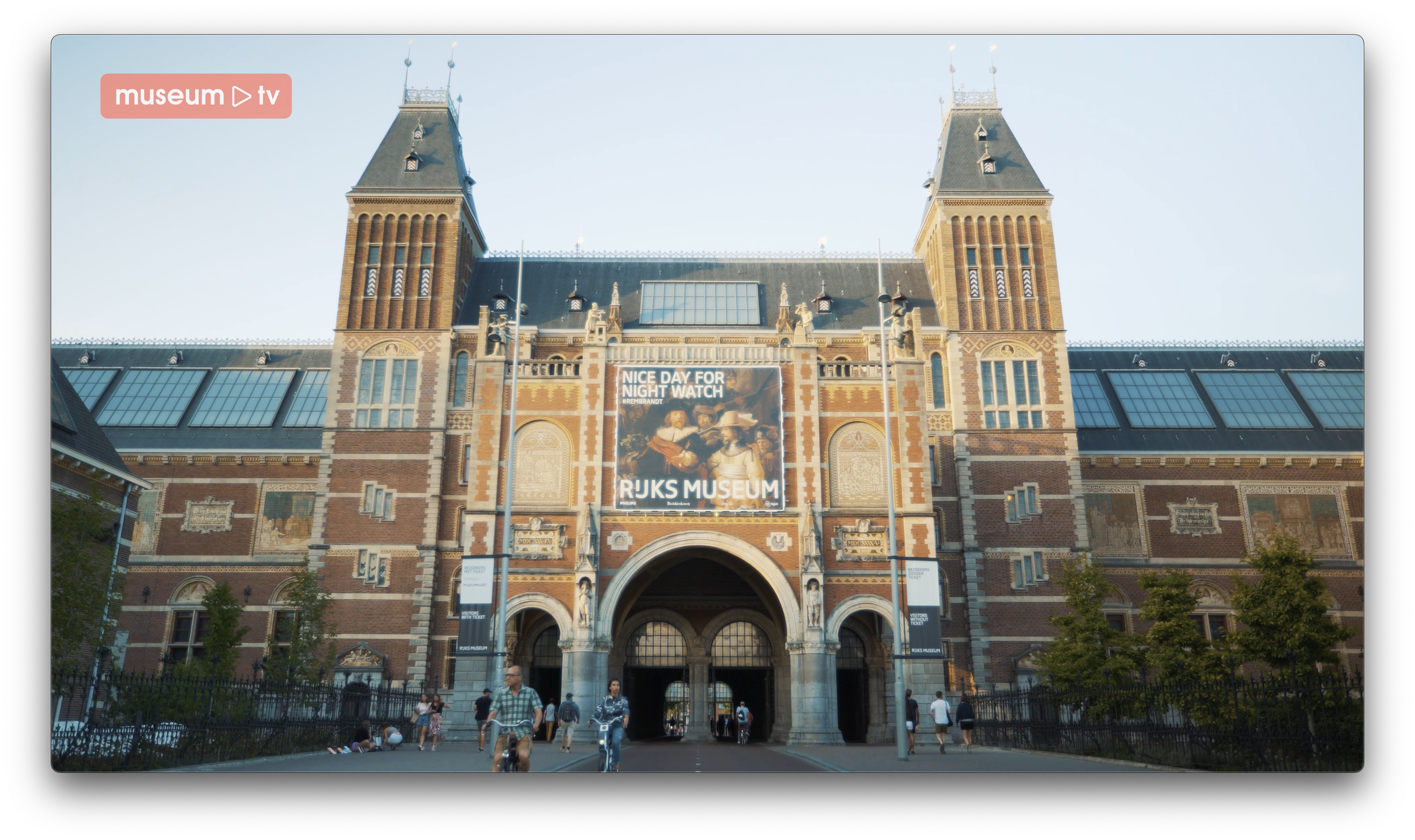 De ingang van het Rijksmuseum in Amsterdam, een groot middeleeuws gebouw met twee hoge torens en een grote poster in het midden. Mensen lopen in en uit de ingang.