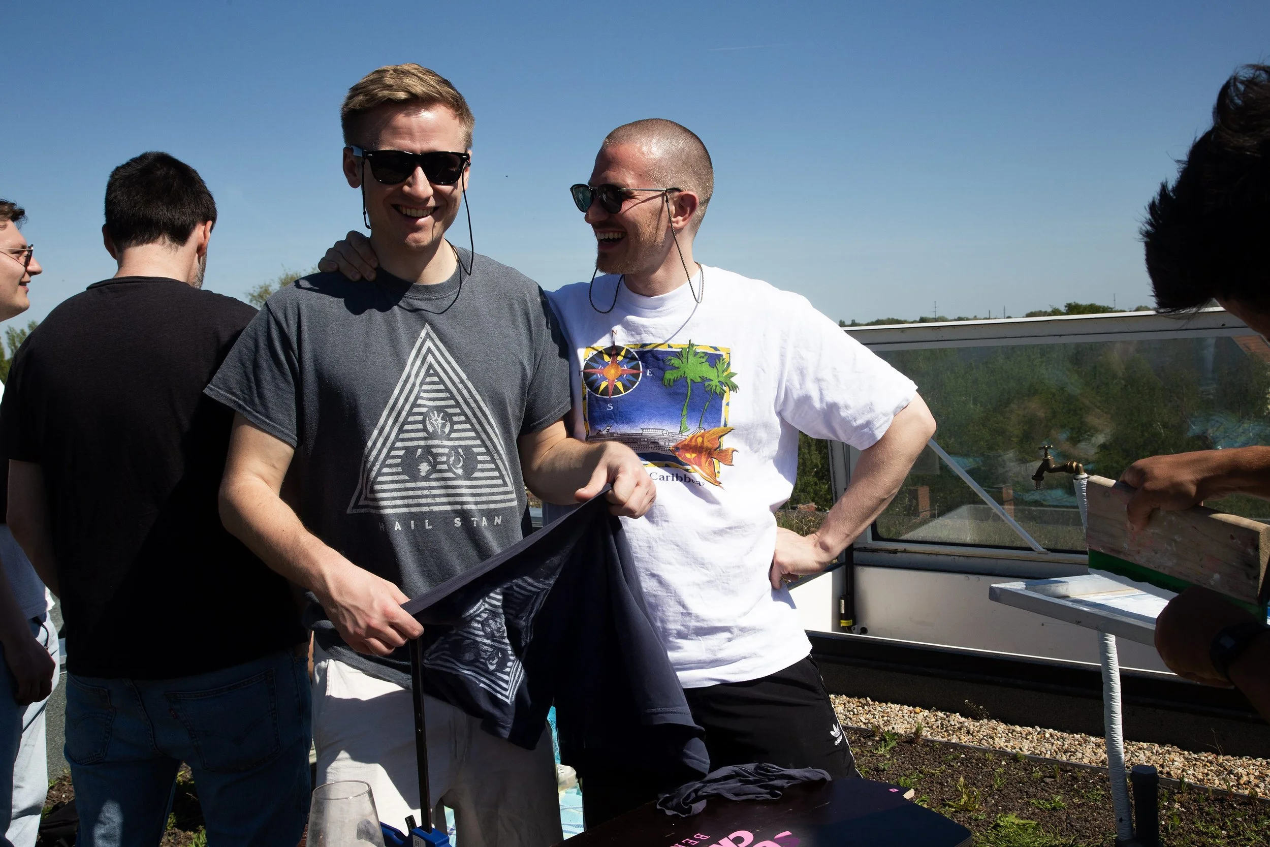 Four young men are outdoors on a sunny day, smiling and talking, with one wearing sunglasses and a grey t-shirt, another with a shaved head and a white t-shirt featuring colorful graphics, and two more in the background.