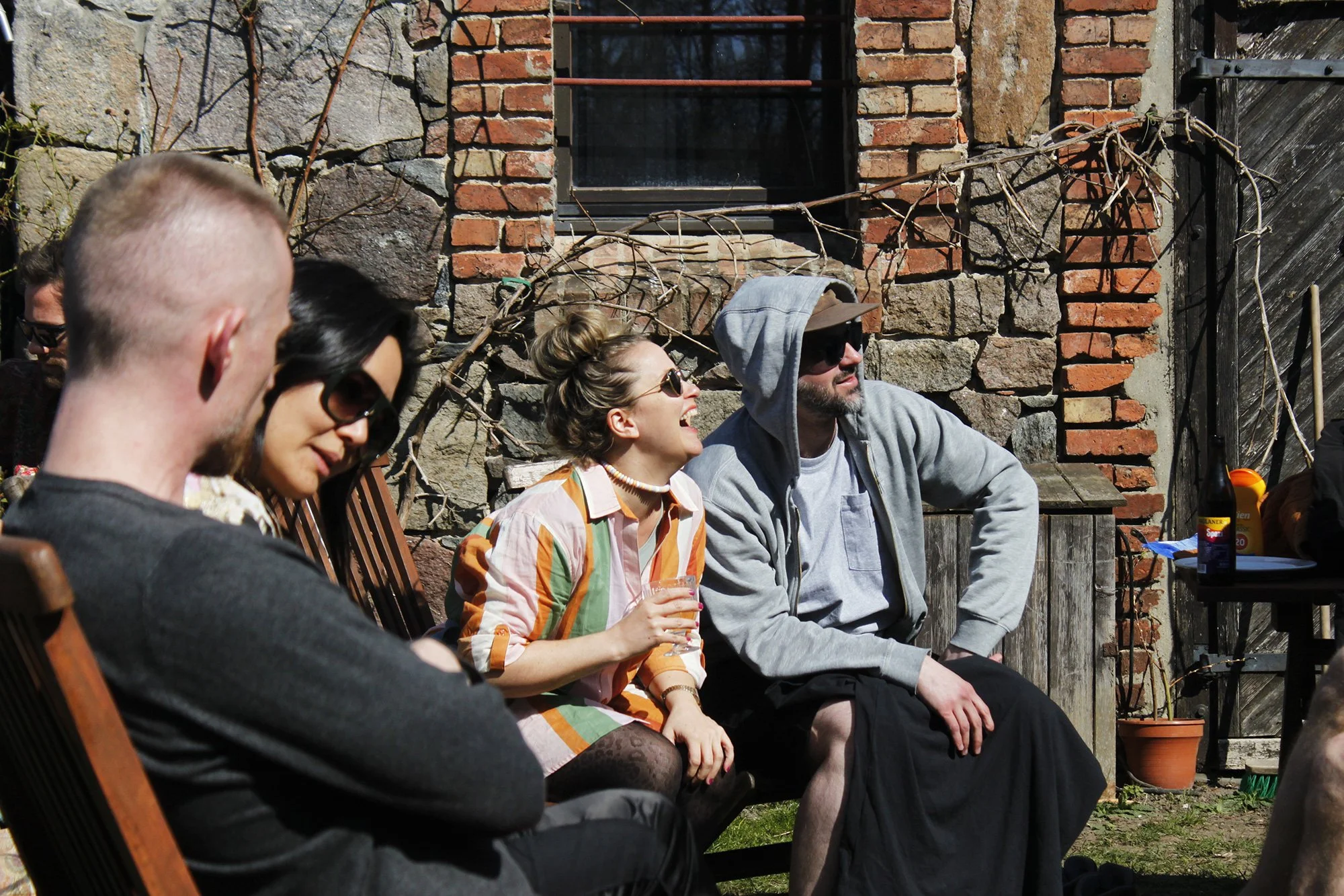 A group of five young adults sitting outdoors against a brick and stone wall, engaged in conversation and laughter, with some wearing sunglasses and casual clothing.