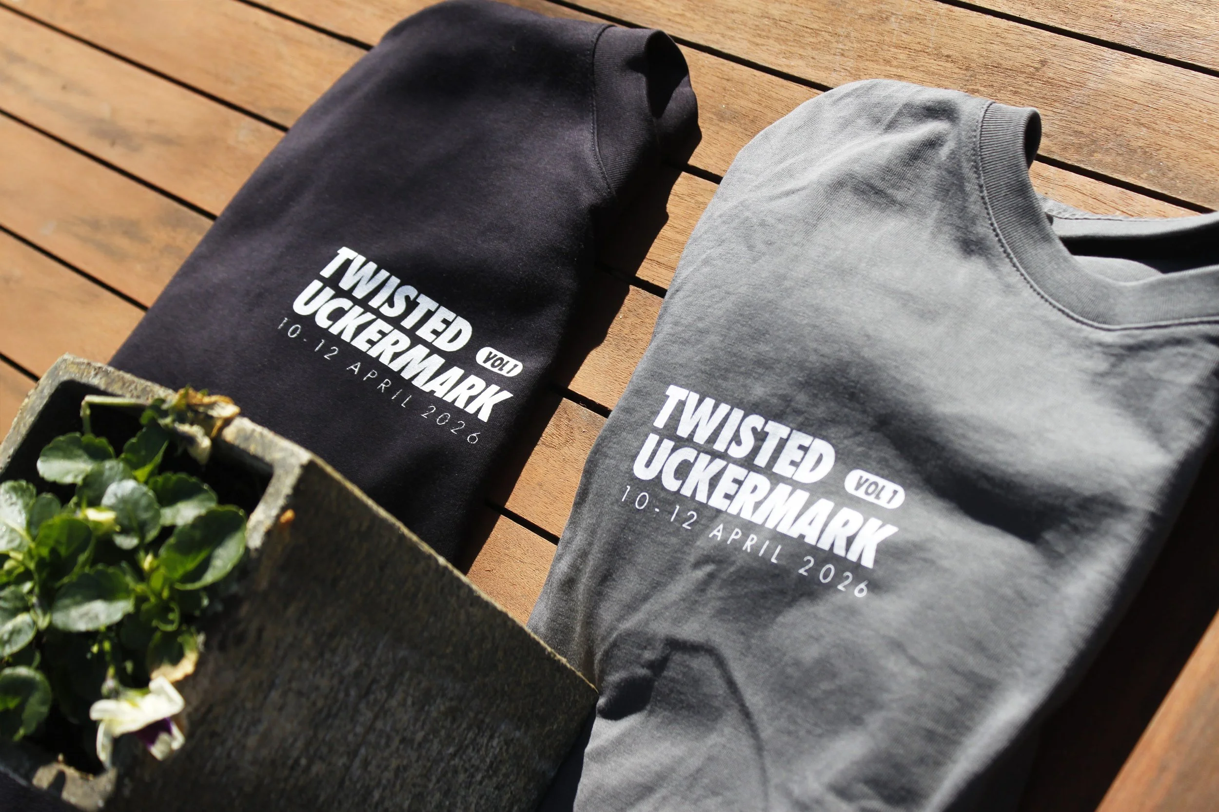 Two folded T-shirts, one black and one gray, with event information printed on them, placed on a wooden surface near a small planter with green plants.