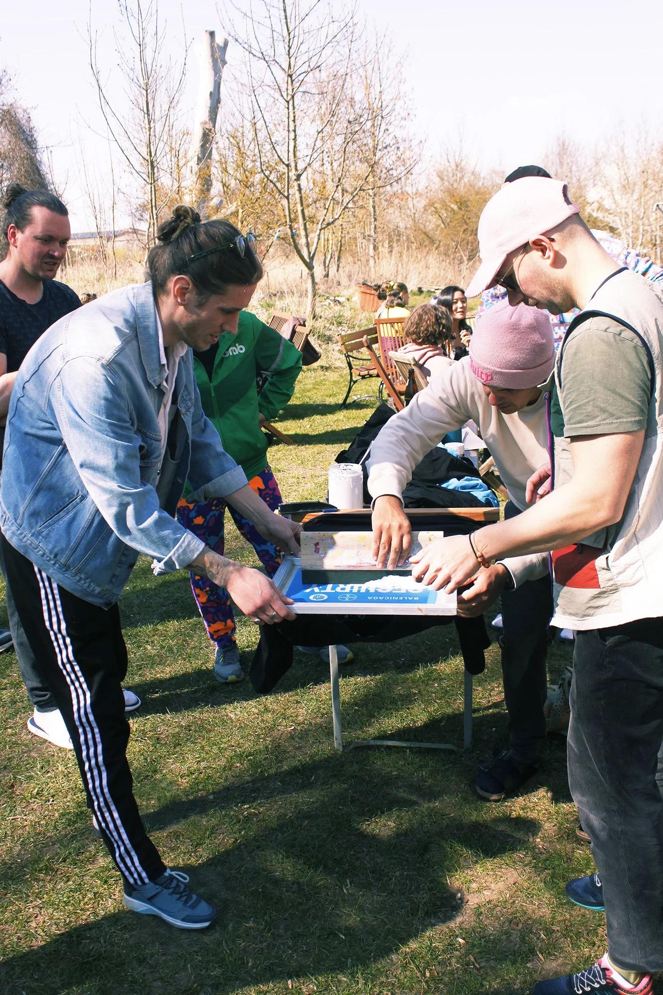 People gathered outdoors, some looking at a table with a game or activity, during a sunny day in a park or garden with trees and benches in the background.