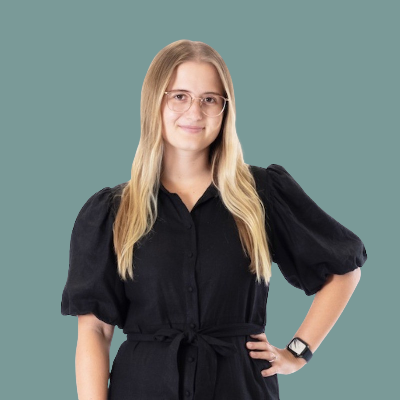 A young woman with long blonde hair, wearing glasses and a black dress, standing against a teal background with one hand on her hip.