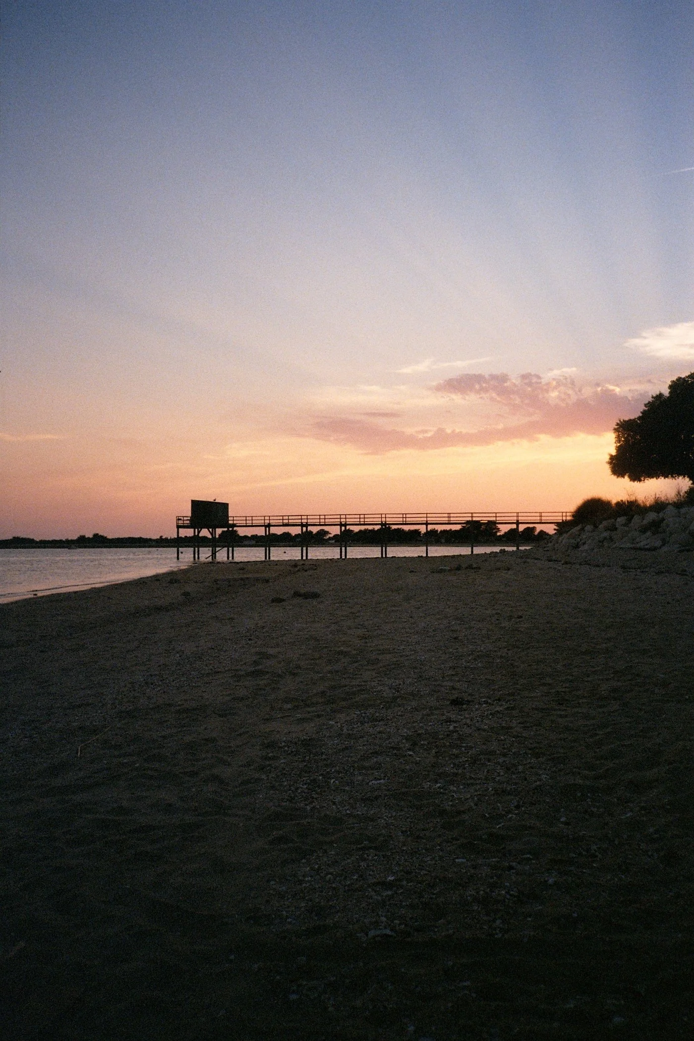Coucher de soleil sur la plage avec un ciel coloré en dégradé, une jetée en bois avec une petite structure sur pilotis et des arbres silhouettés sur la droite.