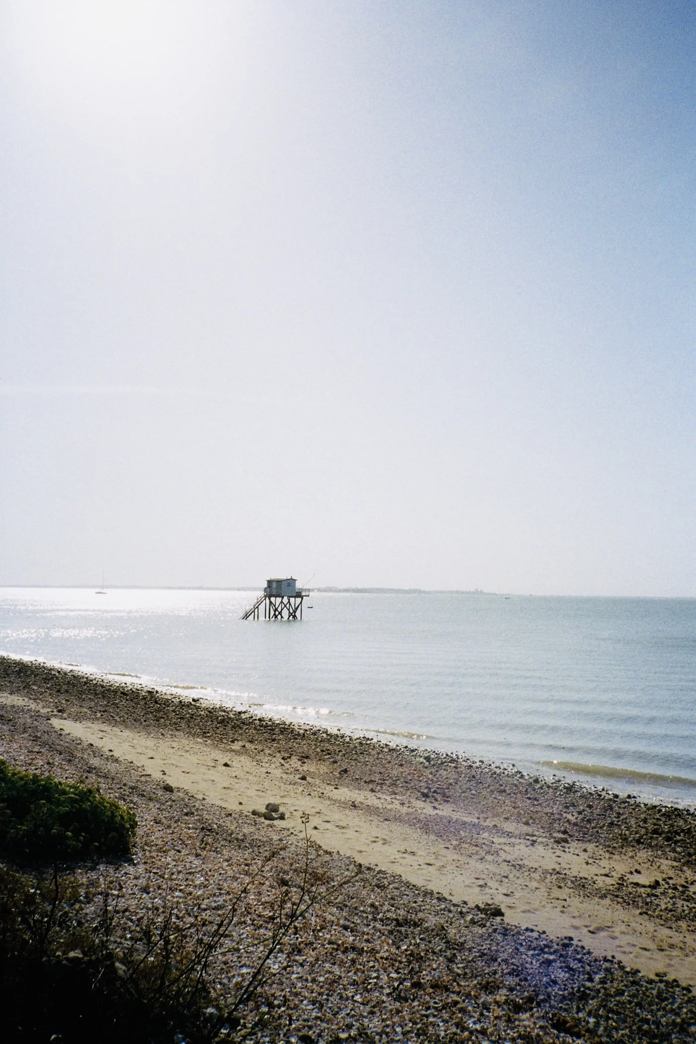 Plage avec des galets, eaux calmes et un cabanon sur pilotis en arrière-plan, ciel clair et horizon visible.