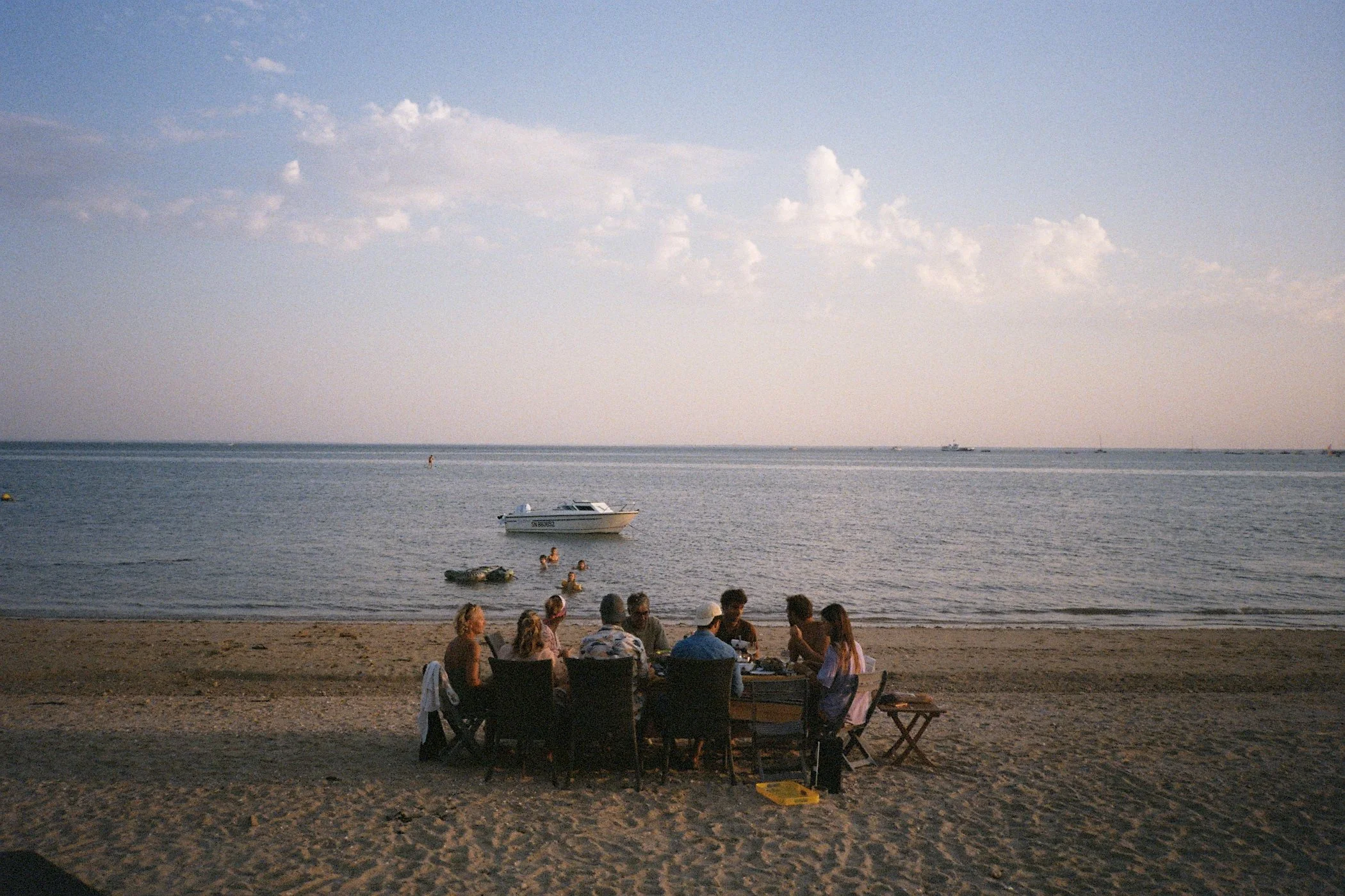 Groupe de personnes assises sur la plage au coucher du soleil, avec un bateau à l'eau et des enfants nageant