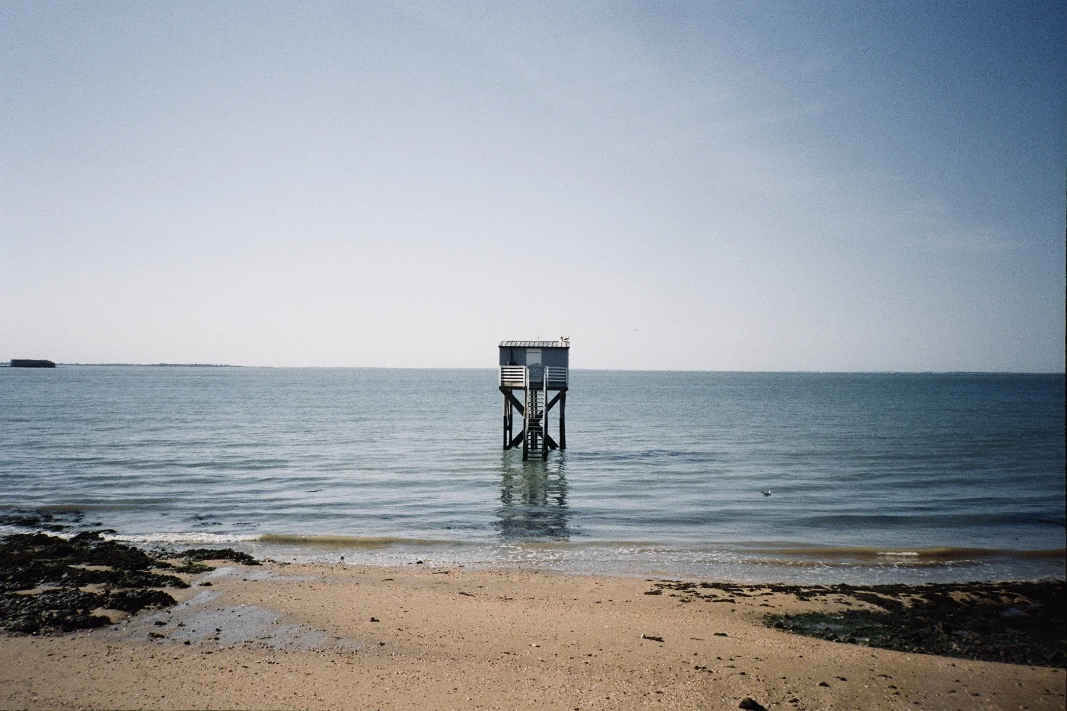 Une cabane de sauvetage en bois sur pilotis au bord de la mer, avec un ciel clair et une plage de sable avec des algues en premier plan.