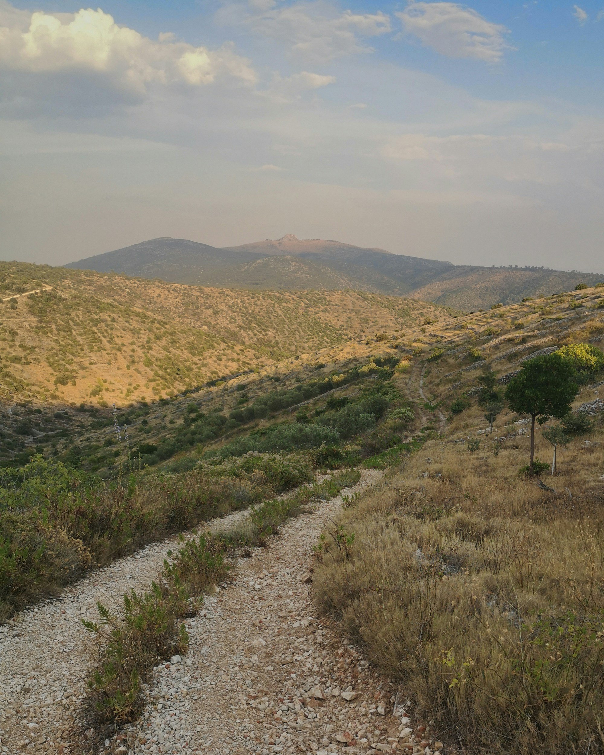 Hiking trail through a dry, rocky landscape with rolling hills and sparse vegetation under a partly cloudy sky.