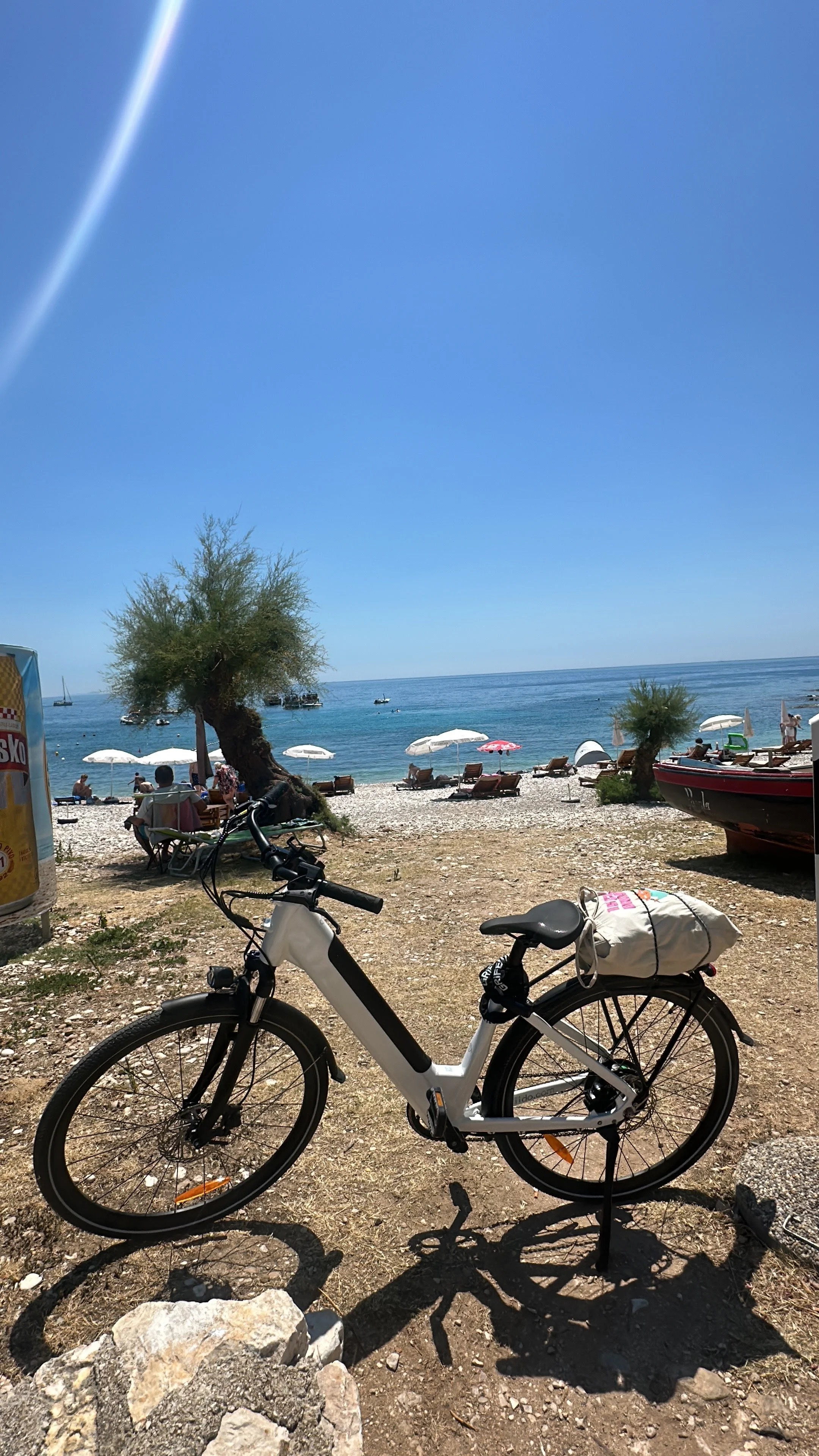 A beach scene with a bicycle in the foreground, white umbrellas, lounge chairs, people relaxing, trees, a boat on the water, and a clear blue sky.