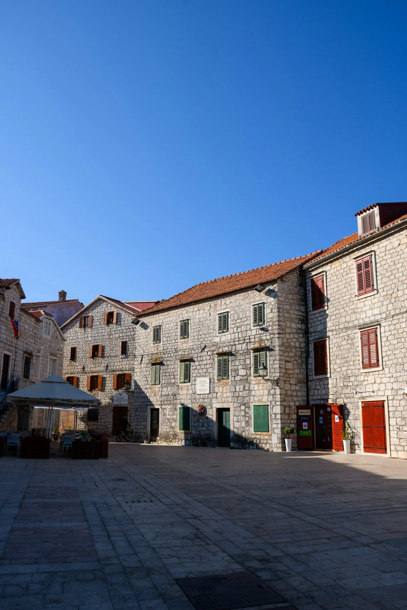 An old stone building with red window shutters and a red-tiled roof, in a town square paved with stone, under a bright blue sky.