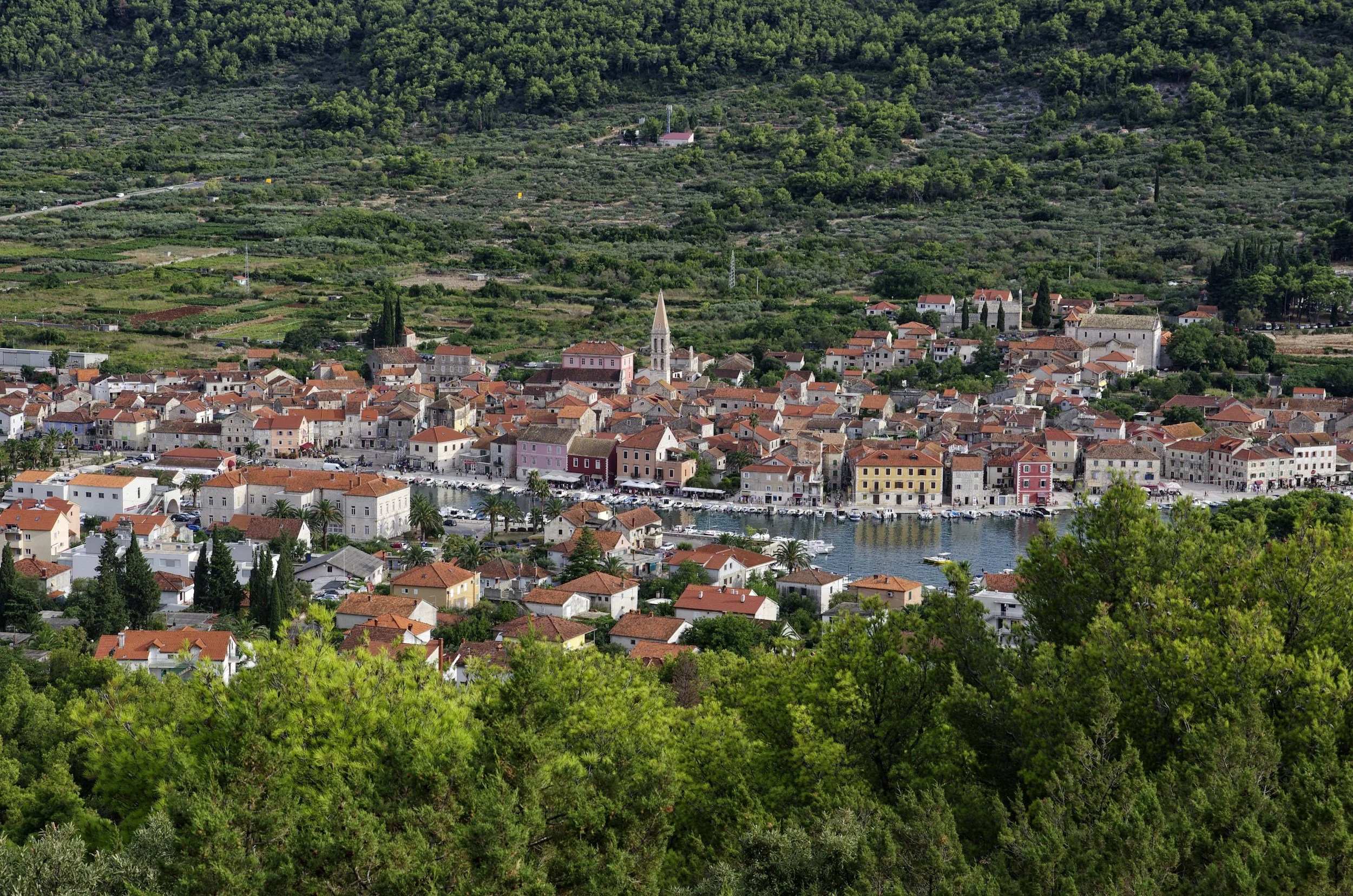 A picturesque view of a coastal town with red-tiled roofs, a harbor filled with boats, and a church with a tall steeple, surrounded by lush green hills and trees.
