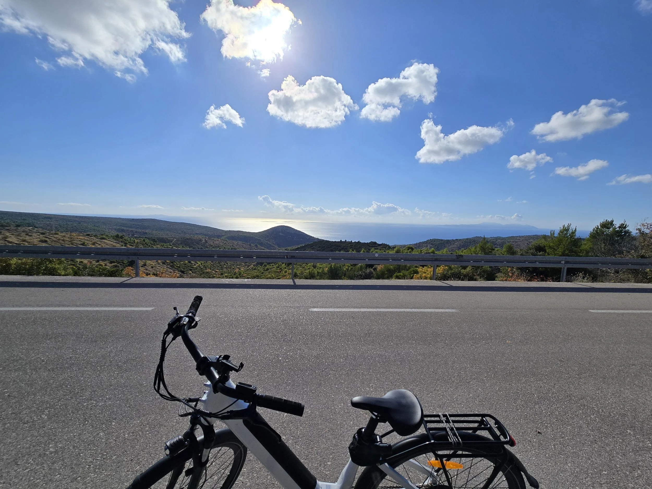 View of a mountainous landscape with the ocean in the distance under a bright blue sky with scattered clouds, taken from a roadside with a bicycle in the foreground.