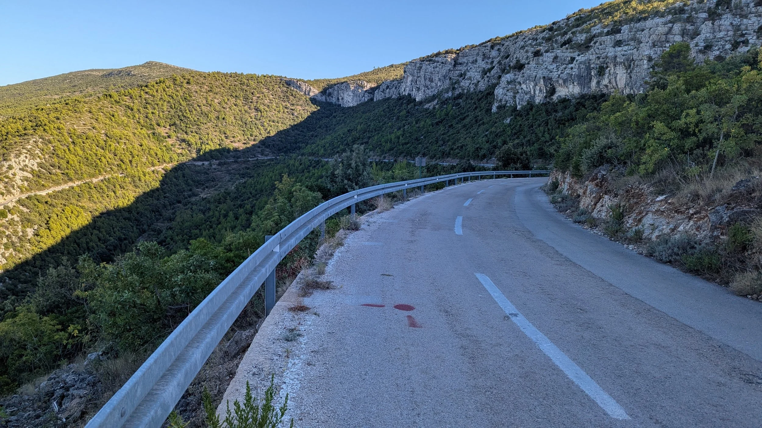 A winding mountain road with a metal guardrail, surrounded by green hills and rocky cliffs, under a clear blue sky.