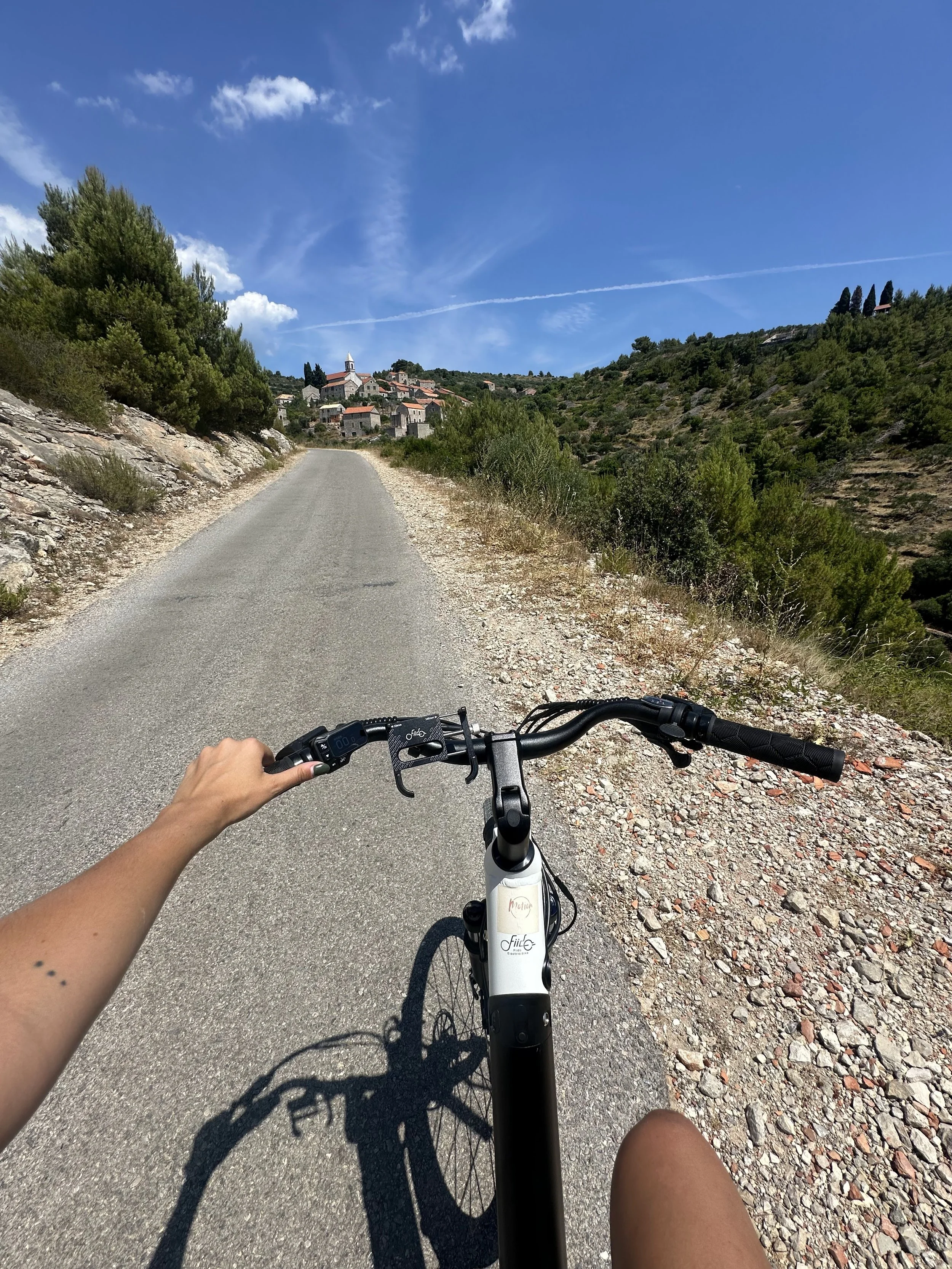 A person riding an electric bike on a narrow paved road through a hillside village with trees and houses, under a bright blue sky with scattered clouds.