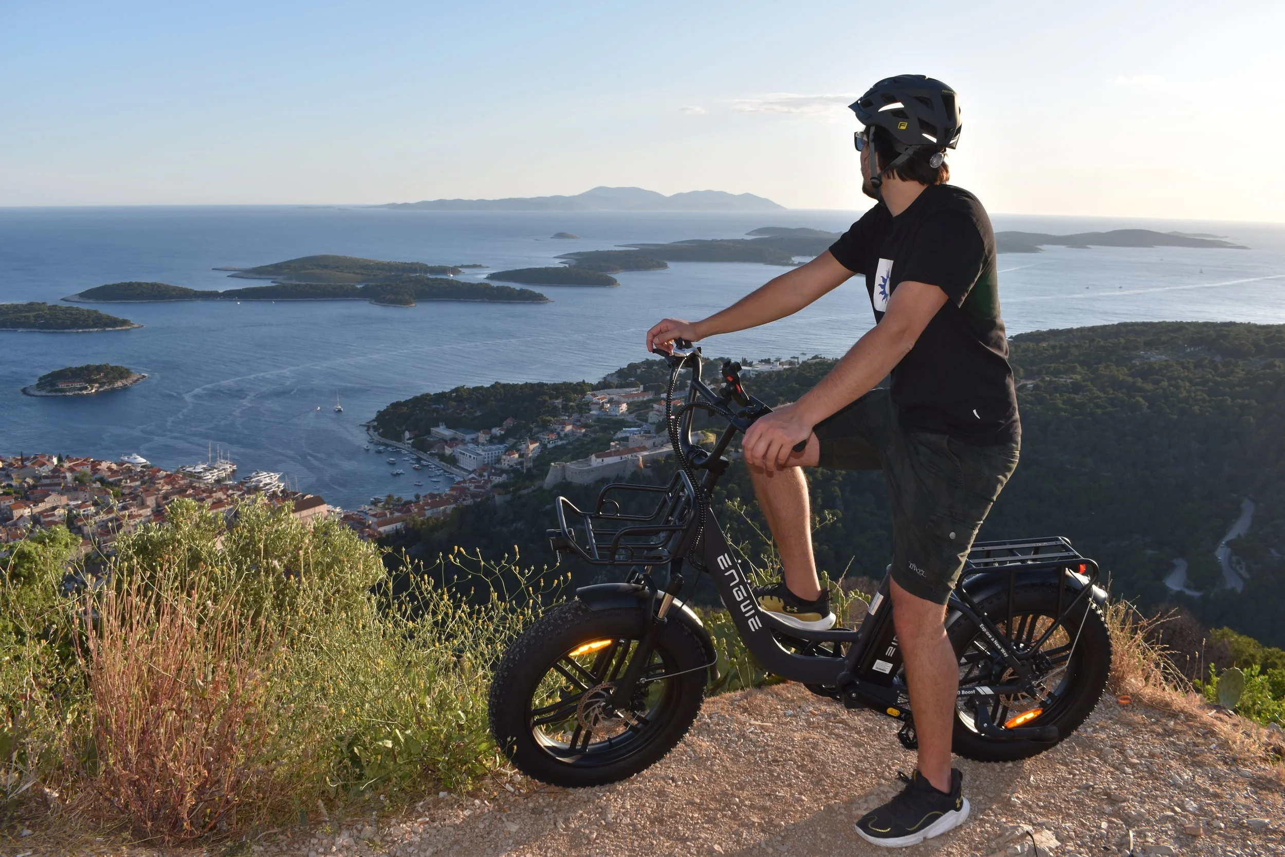 A person wearing a black helmet, black t-shirt, black shorts, and sunglasses riding a black bicycle on a mountain trail overlooking a coastal town and islands in the ocean during sunset.