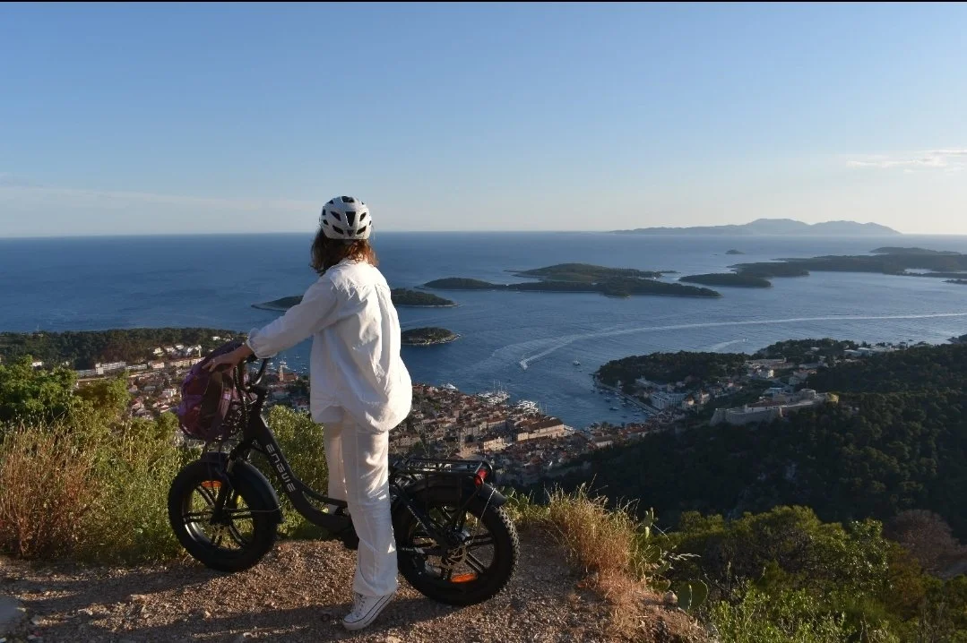 A woman in white clothing and a helmet standing next to an electric bicycle on a hillside overlooking a coastal town with islands and water in the background.