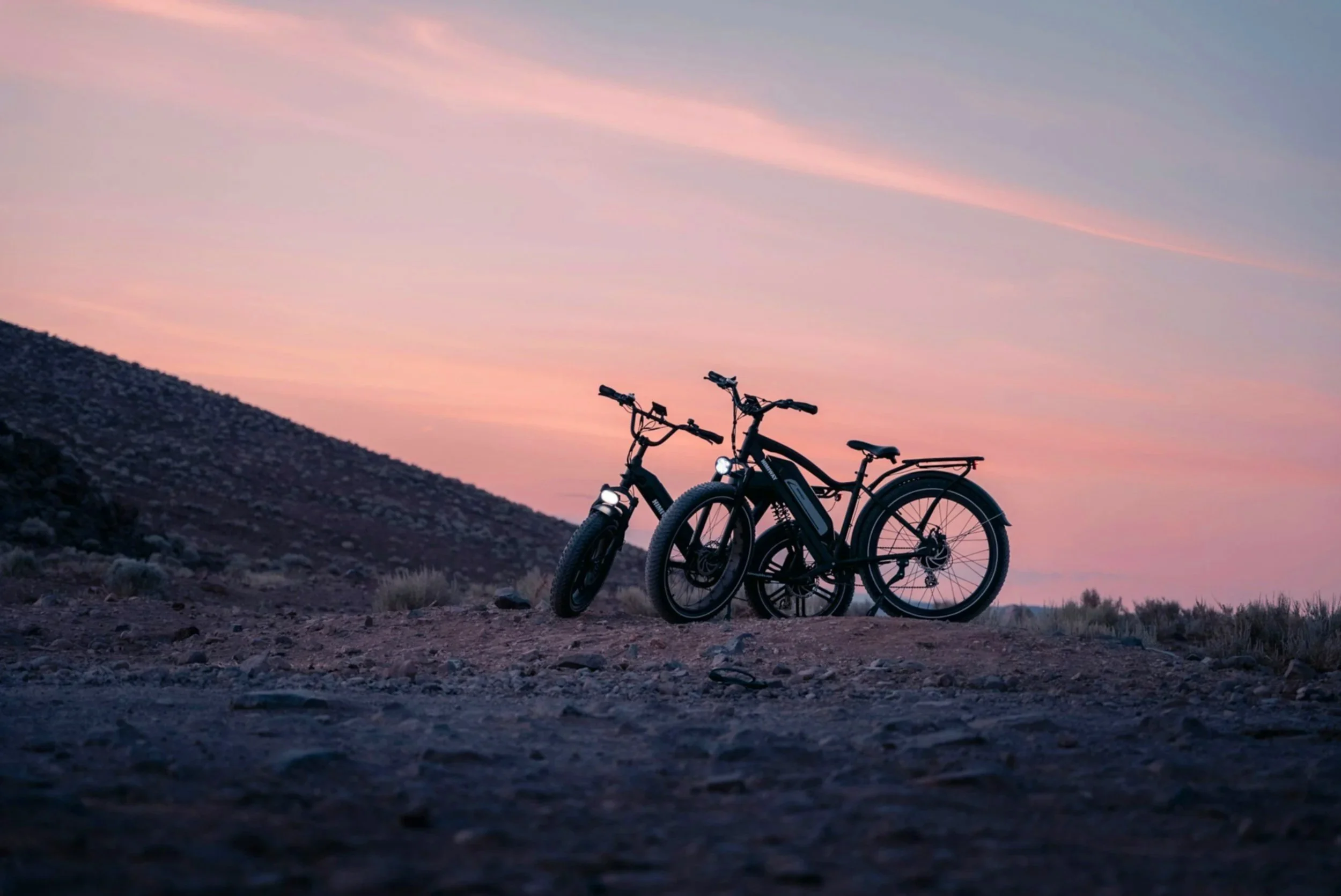 Two bicycles with front lights parked on a dirt ground during sunset, with a pastel pink and blue sky and a rocky hillside in the background.