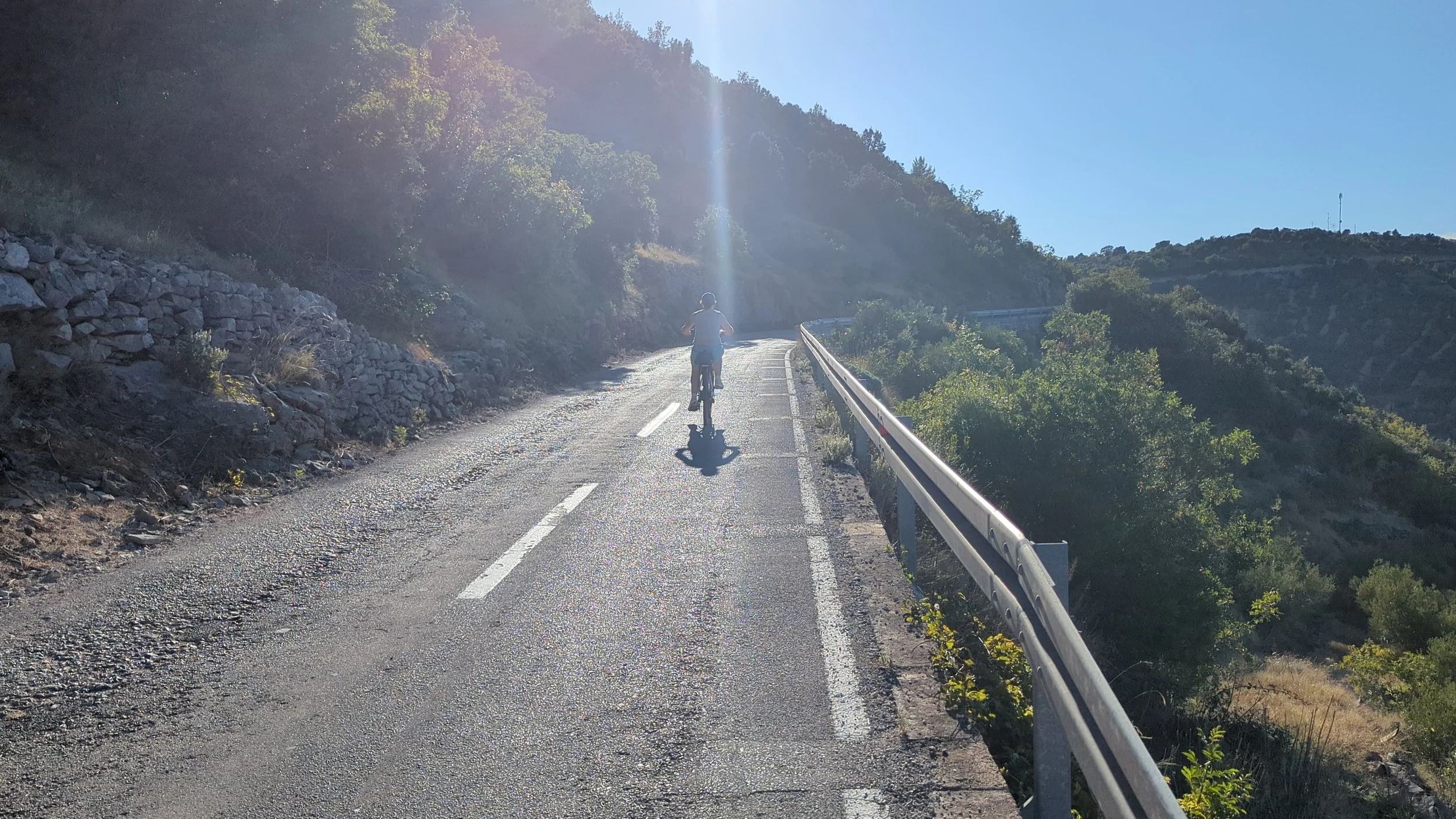A person riding a bicycle on a mountain road in the daytime, with a guardrail on the right and green hills in the background under a clear blue sky.
