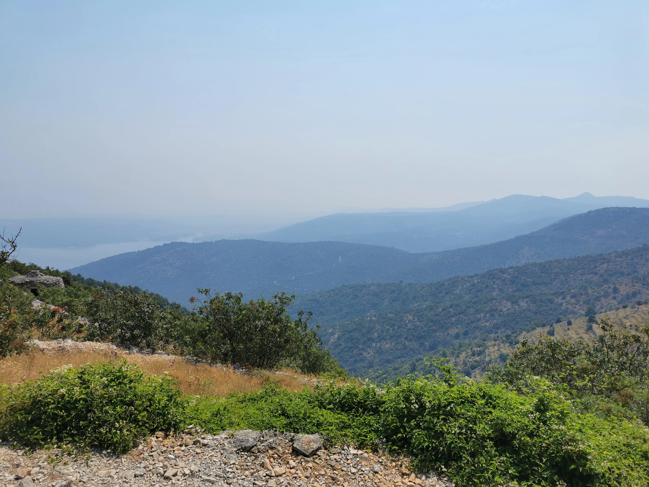 Scenic view of layered mountains with lush green trees in the foreground, a body of water in the distance, and a clear blue sky above.
