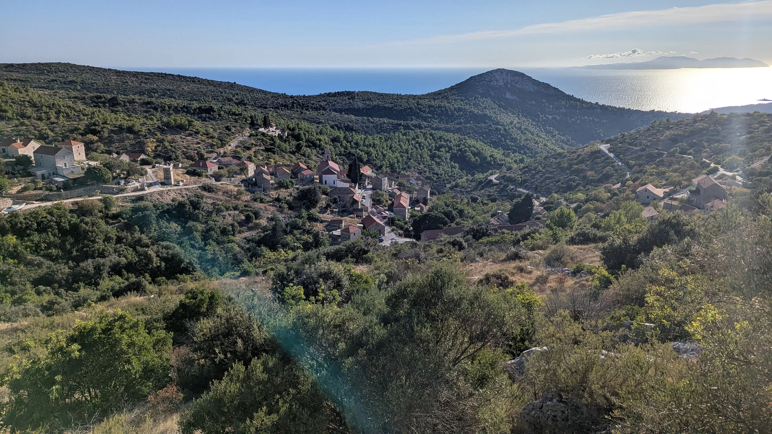Scenic view of a hillside village with houses and church, surrounded by green trees, overlooking the sea with a mountainous island in the distance, under a partly cloudy sky.