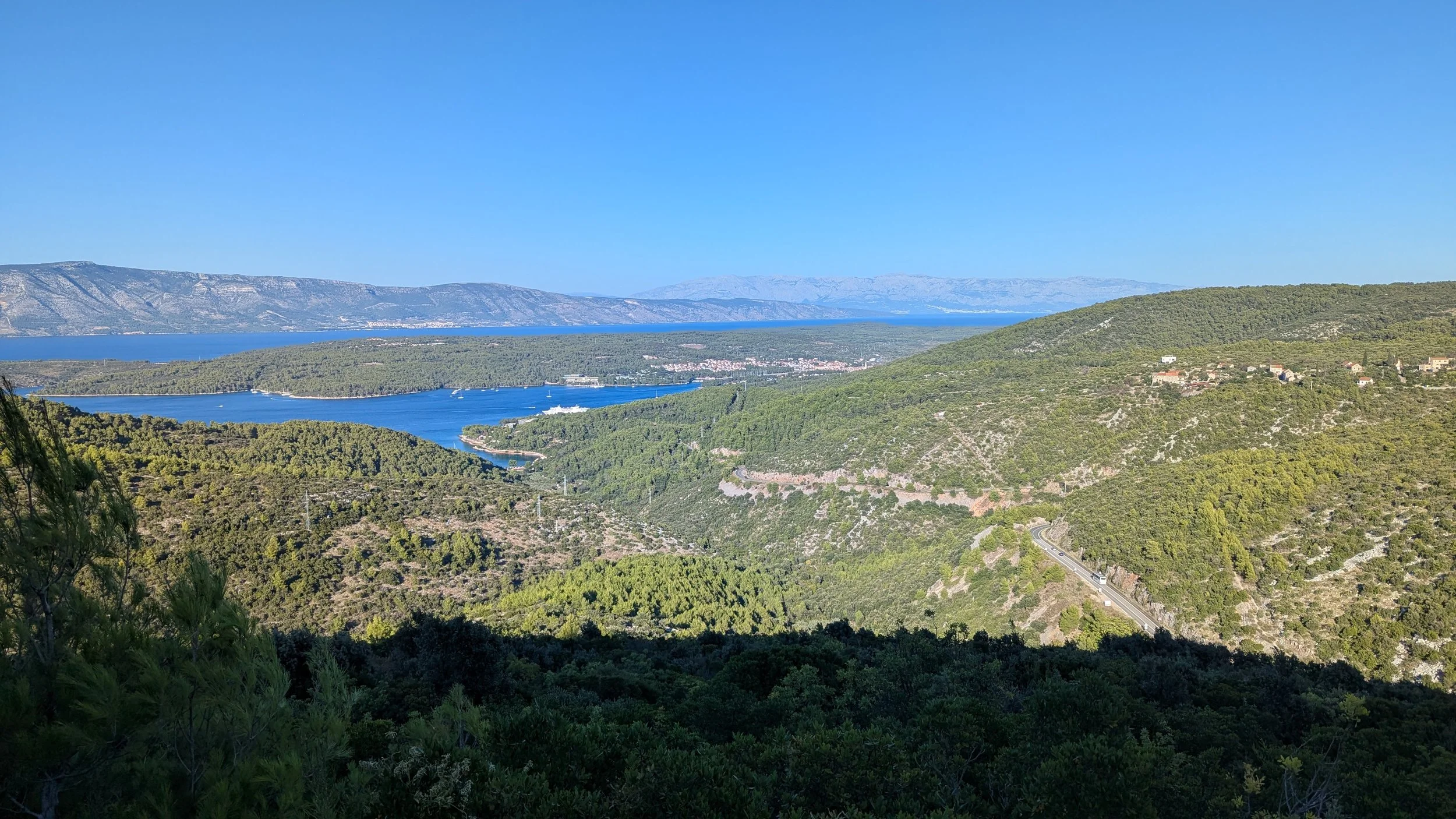 Scenic view of a large body of blue water surrounded by green hills and mountains under a clear blue sky, with some small white boats on the water.