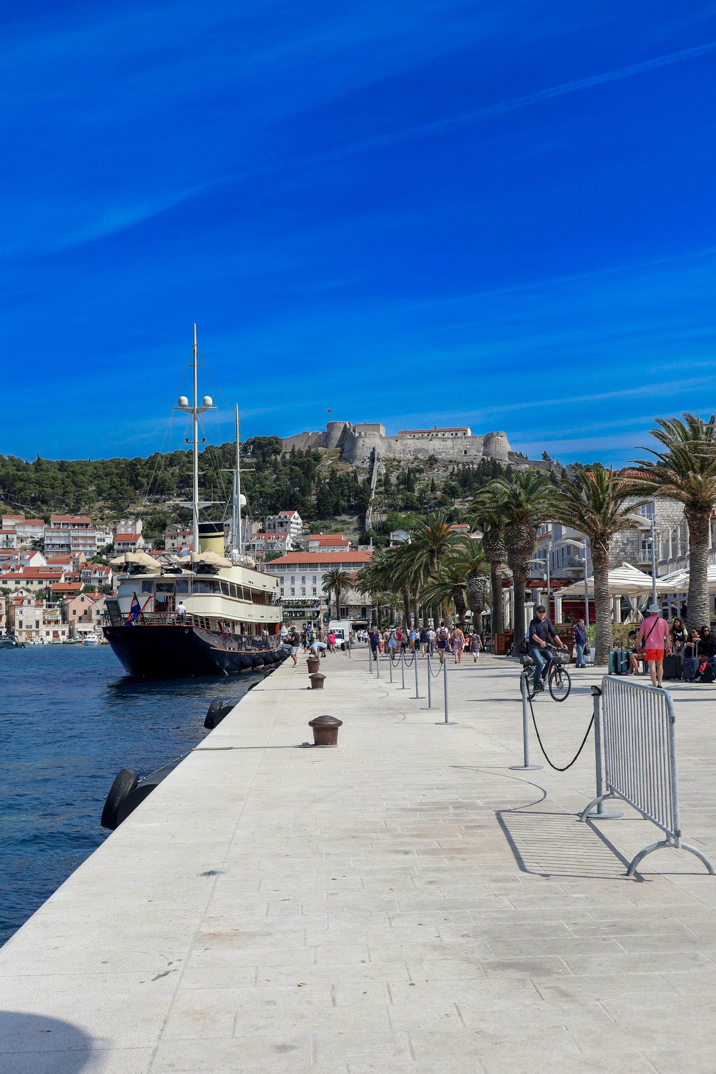 A harbor promenade with people walking, cycling, and relaxing, a boat docked along the quay, palm trees, a hill with a historic fortress or castle on top, and a bright blue sky with wispy clouds.