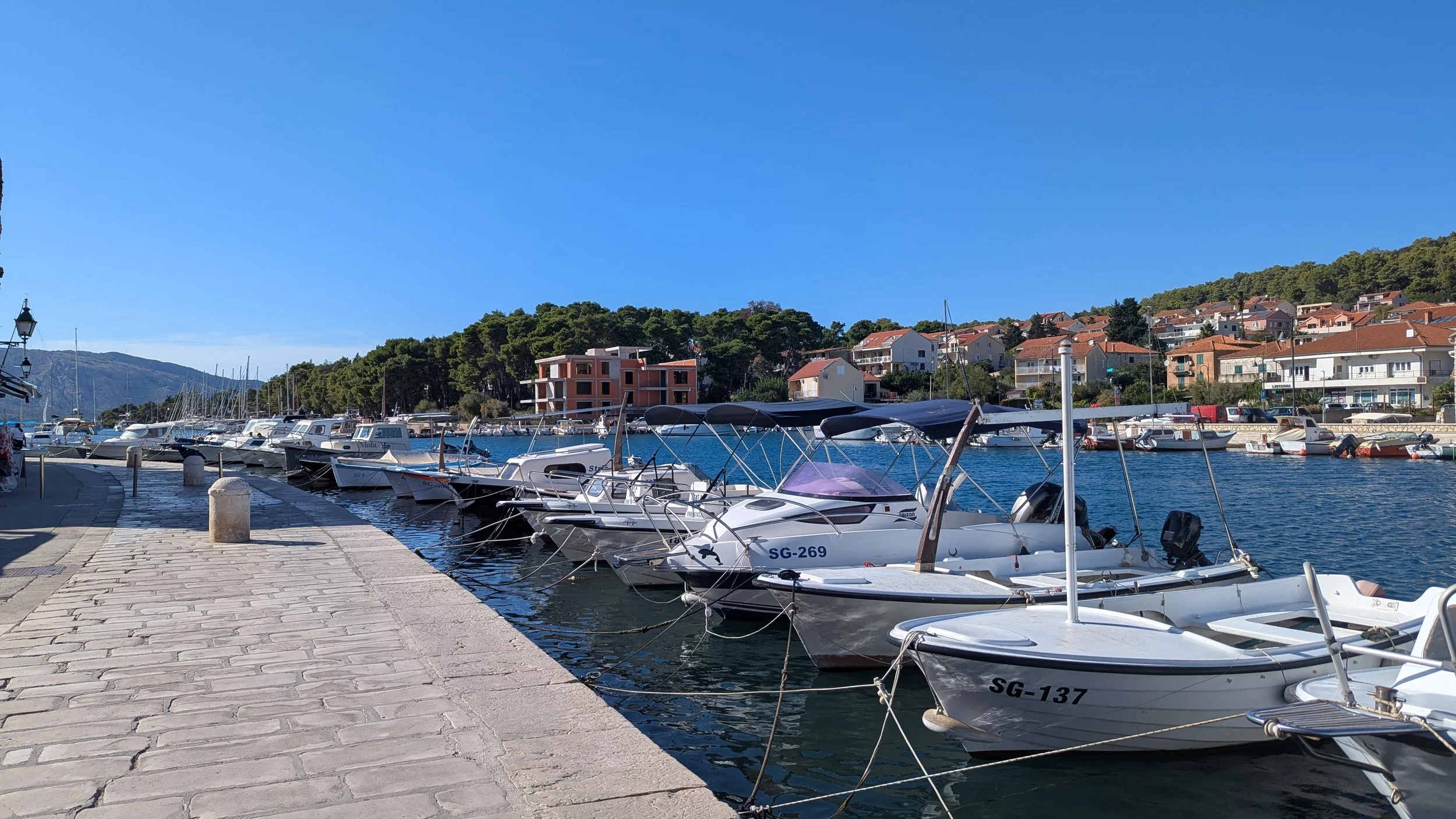 A marina with boats docked along a paved walkway under a clear blue sky, with colorful hillside houses and green trees in the background.