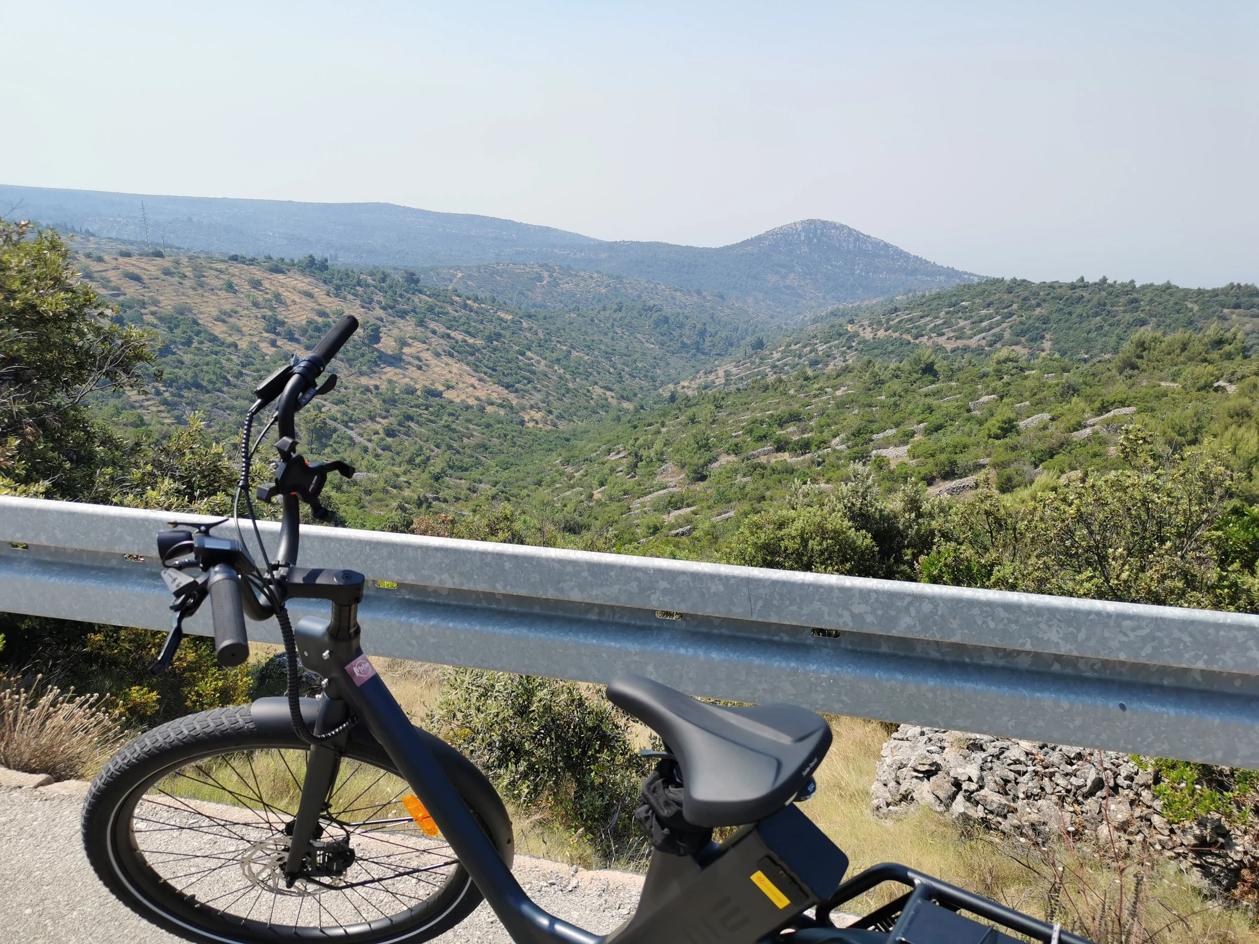 View of green rolling hills with a mountain in the distance, bike handlebars and a bicycle tire in the foreground, metal guardrail along the road, clear sky.
