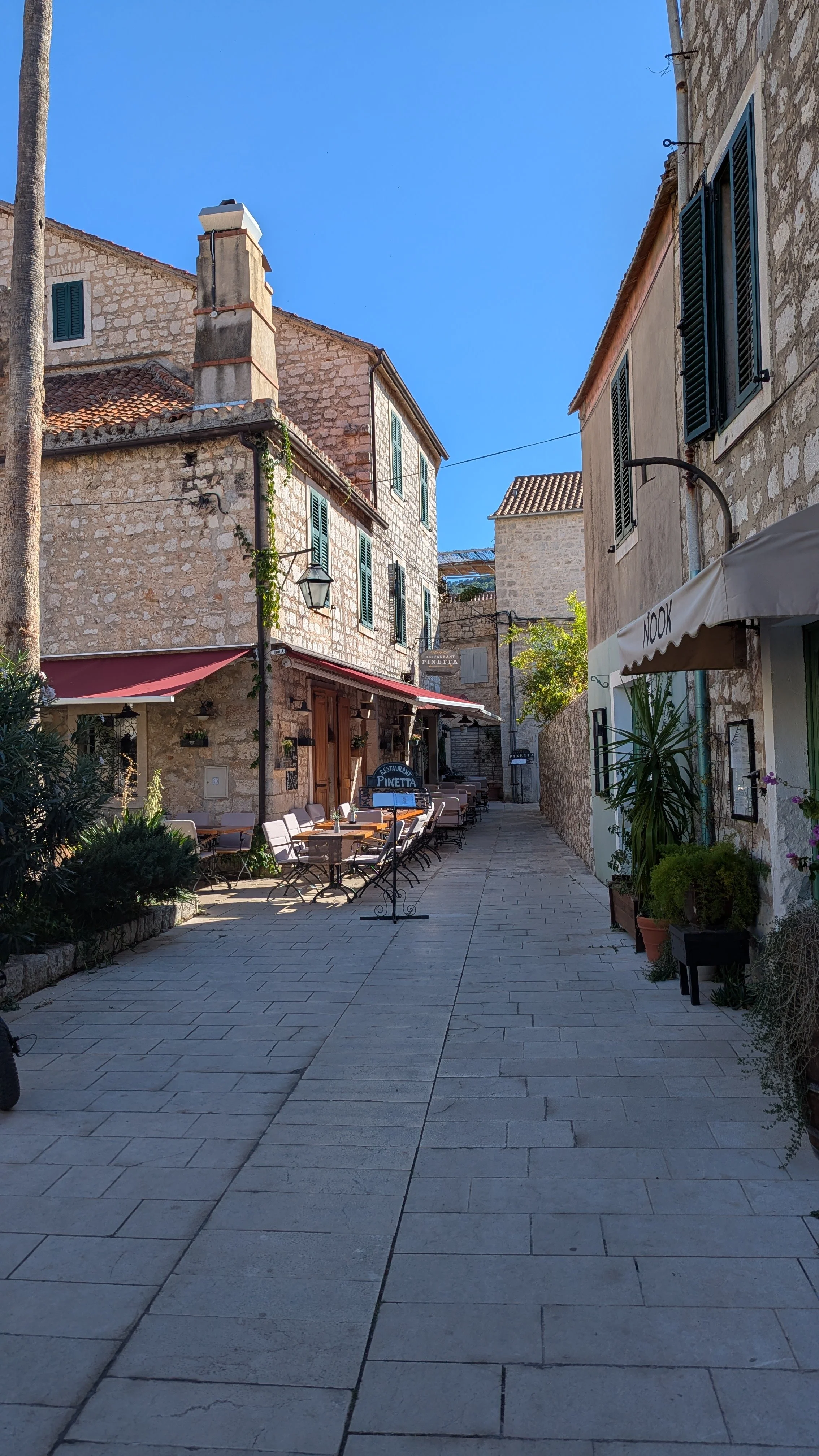 A narrow street in a European town with outdoor restaurant seating, stone buildings with green shutters, and a clear blue sky.