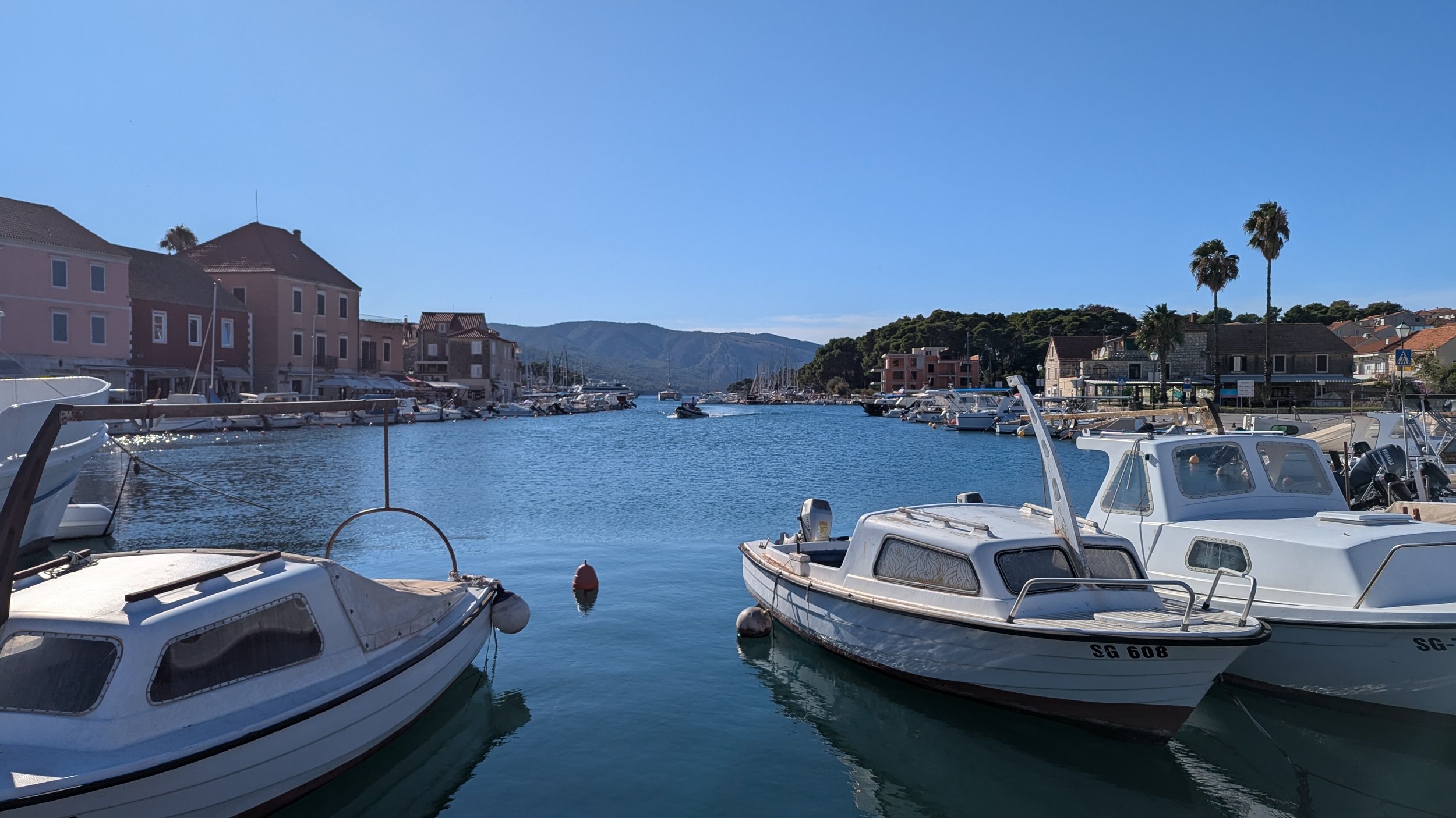 Boats docked in a marina with pastel-colored buildings, mountains in the background, and a clear blue sky.